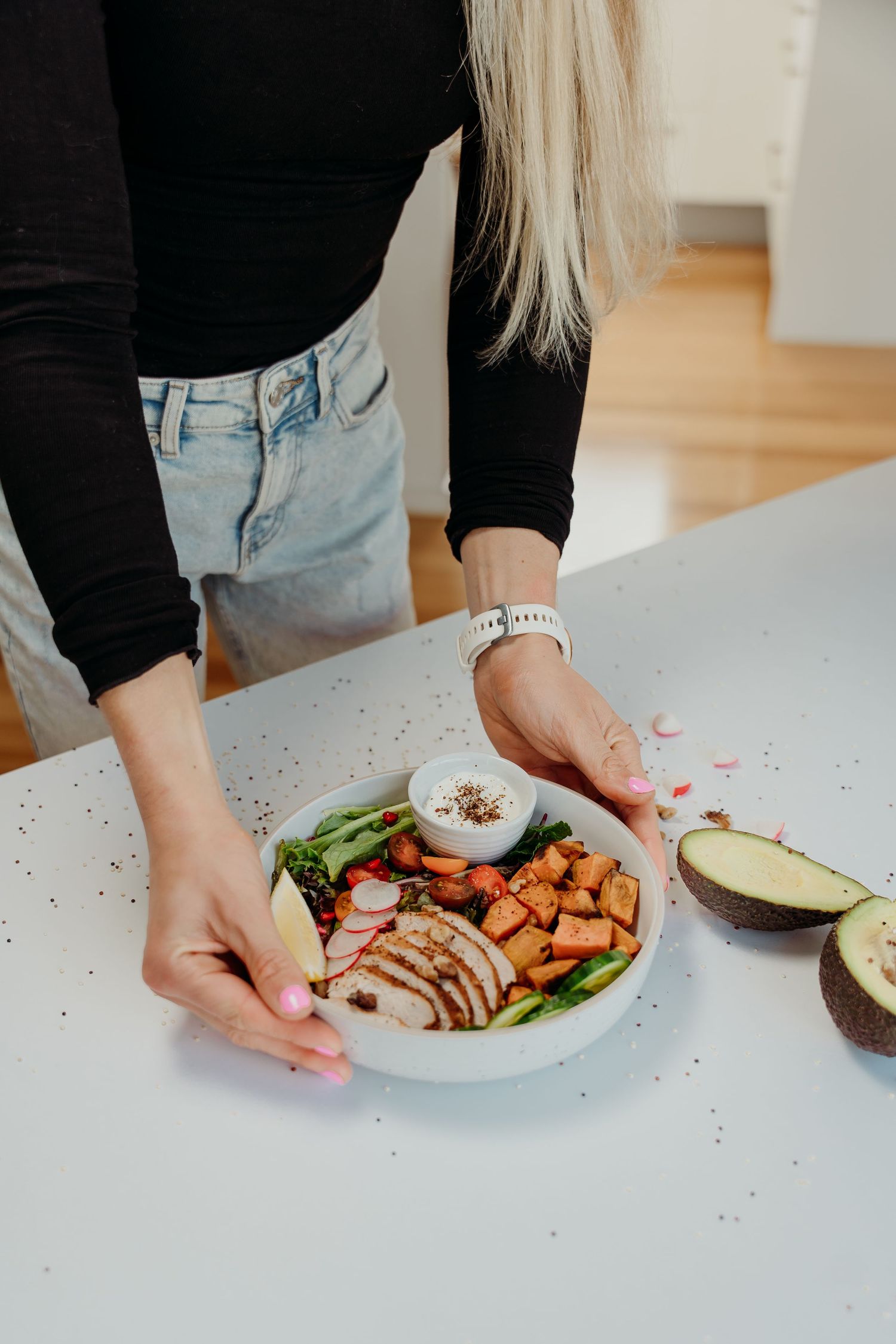Blonde woman arranging a healthy Buddha bowl with chicken, sweet potato, avocado, and greens on a white kitchen counter.