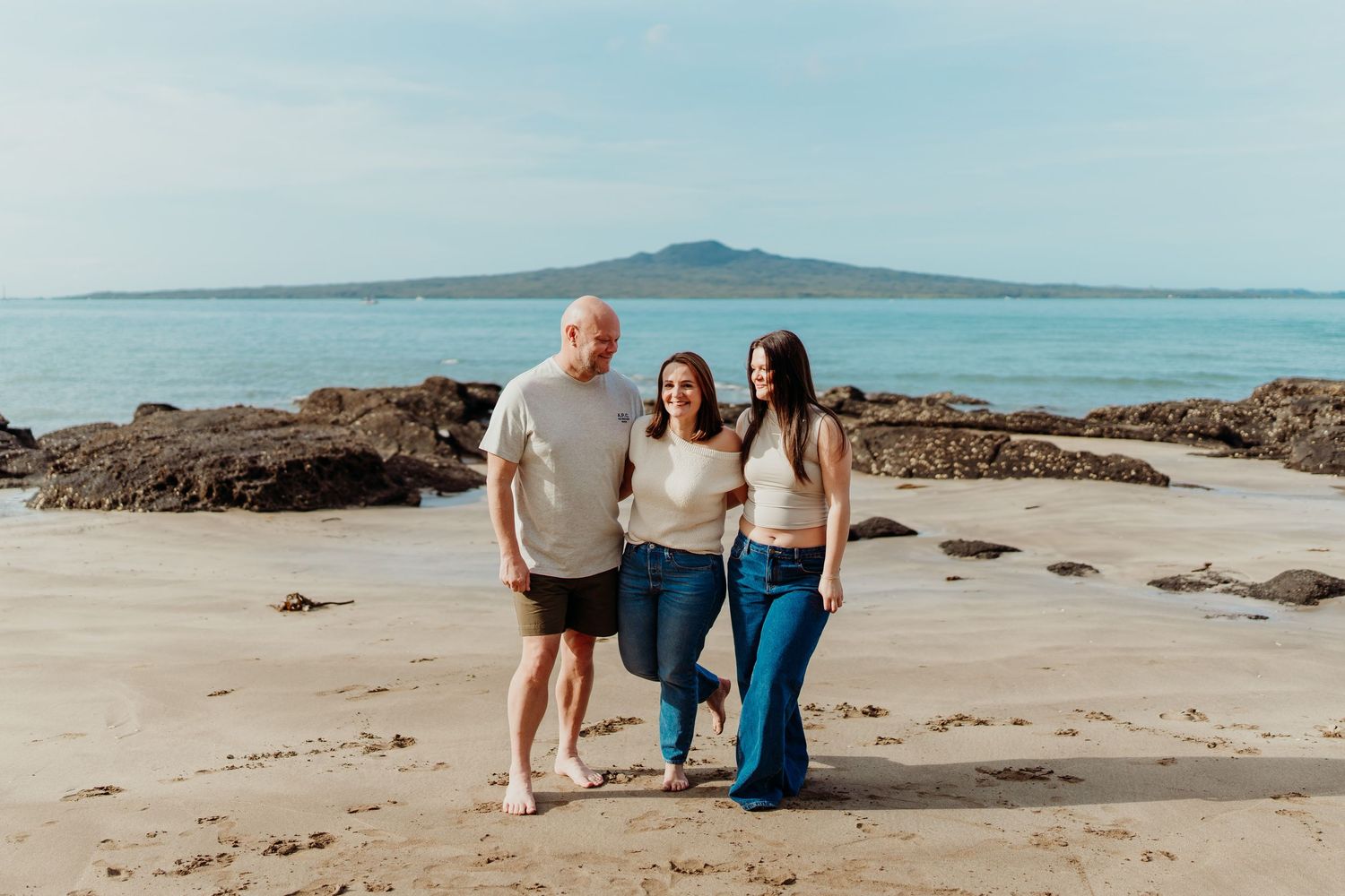 Three people smiling on a sandy beach with a volcanic island visible across calm blue waters in the background.