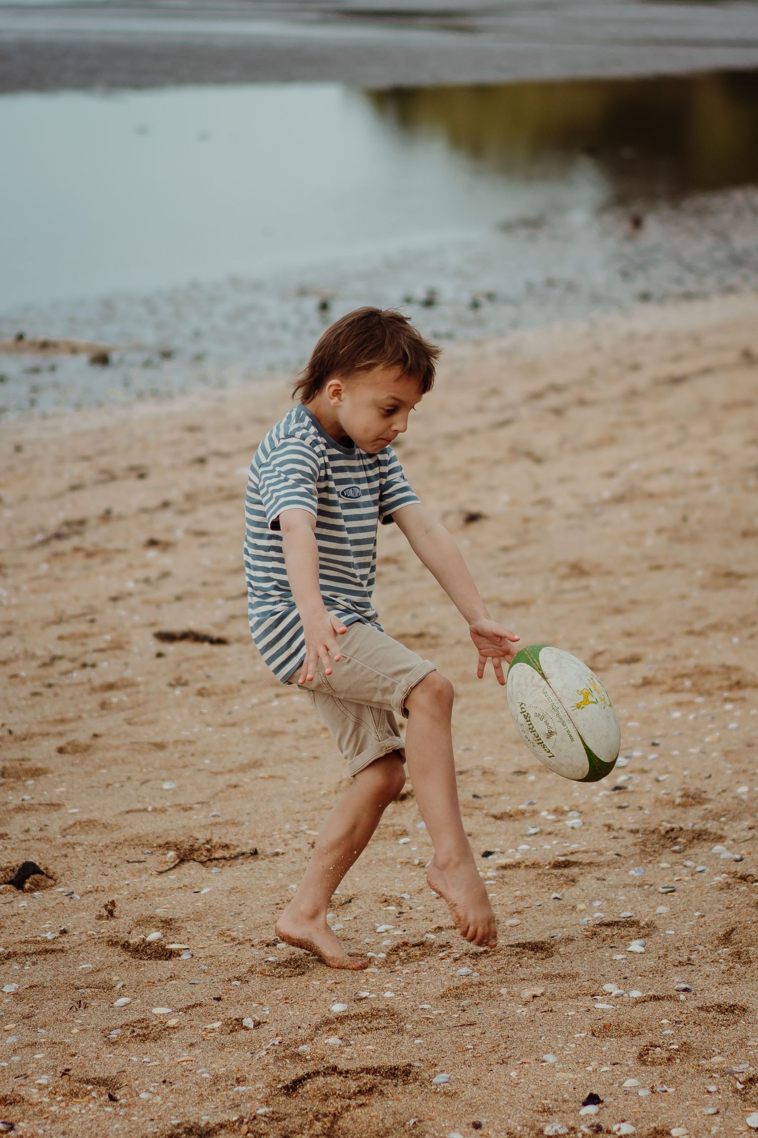 A child in a striped shirt plays on a sandy beach, bending down to pick something up from the shore.