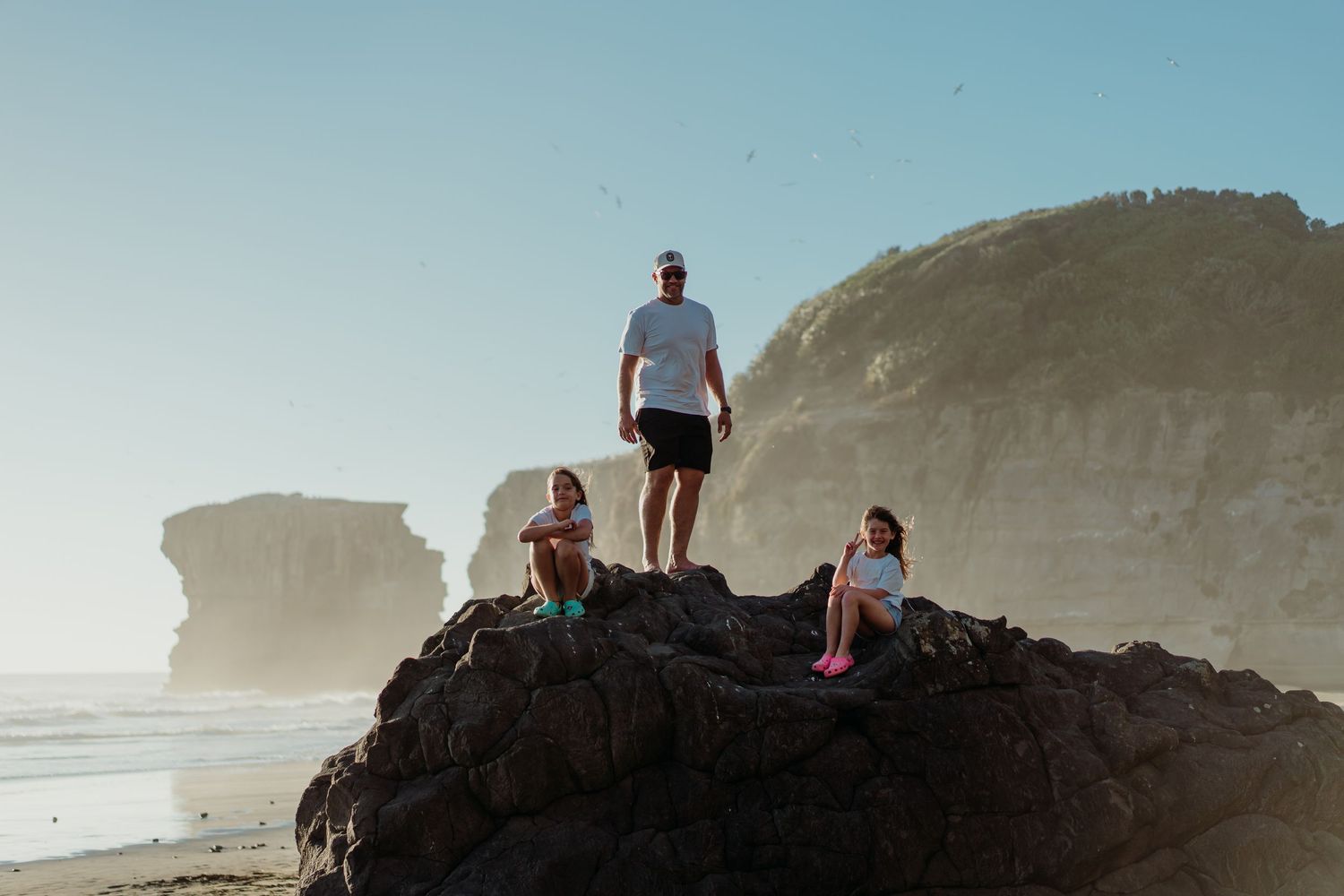 A man and two children stand on a rocky outcrop at a scenic beach with dramatic sea stacks in the background.