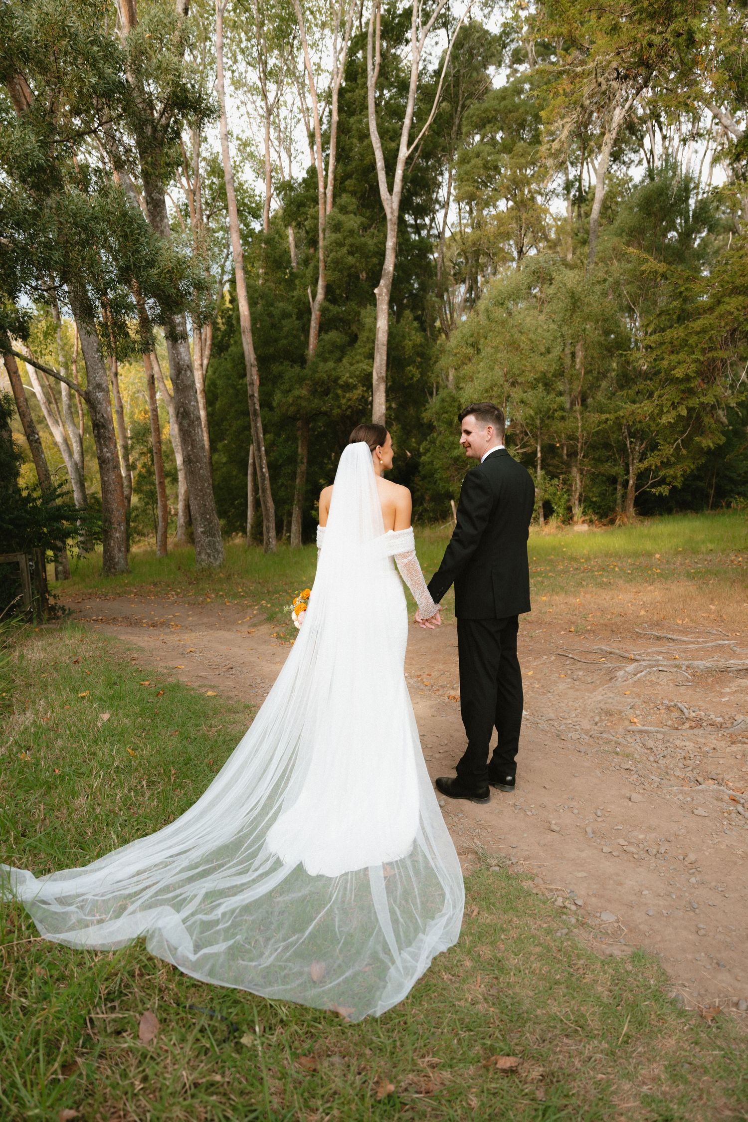 A flowing white wedding dress train trails behind a couple walking on a dirt path through eucalyptus trees.