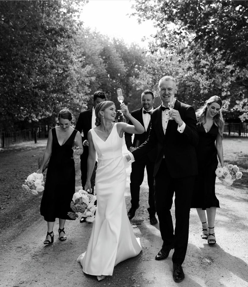 A wedding party walks along a tree-lined path in black and white, creating a classic processional scene.