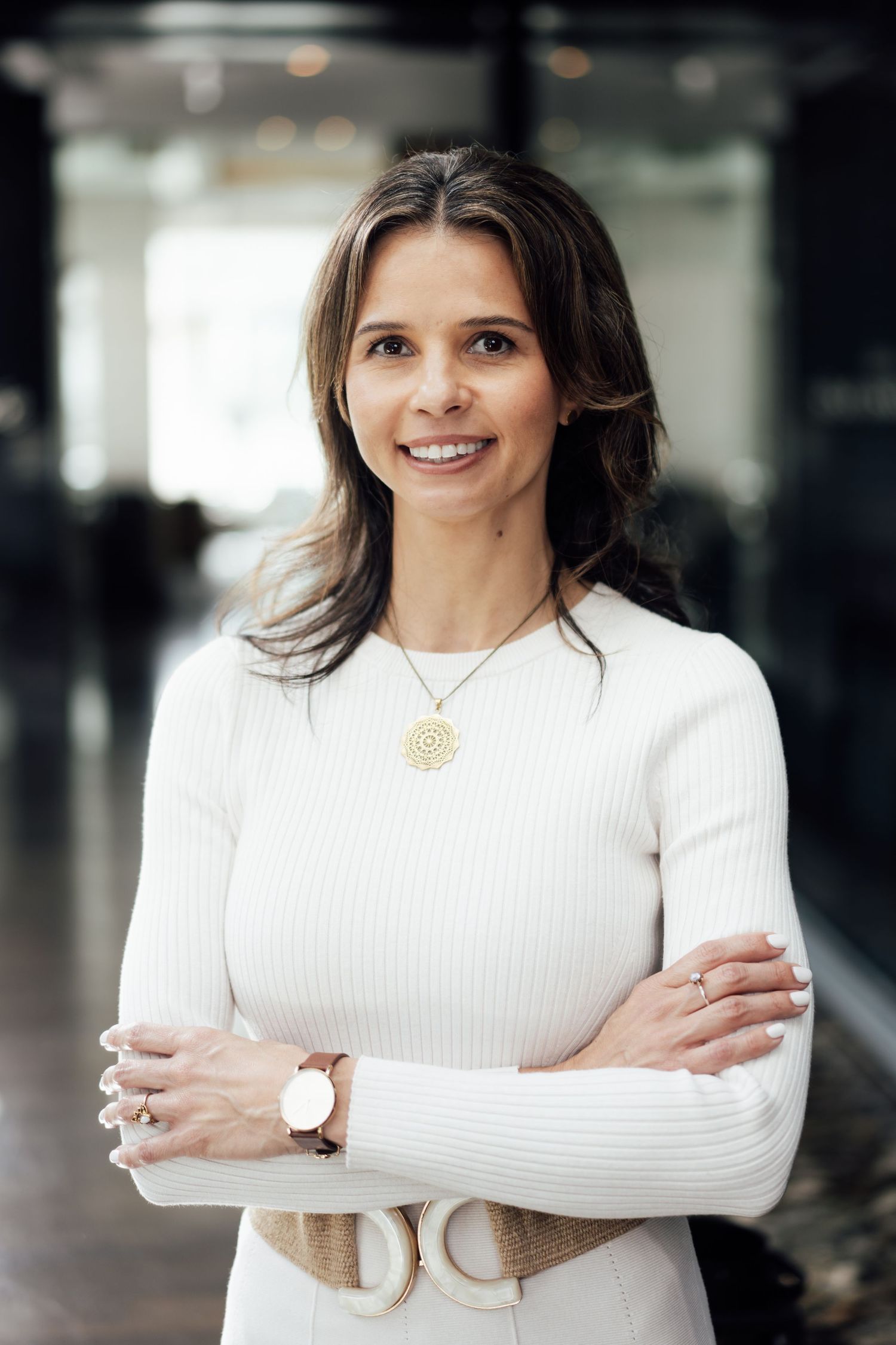 Professional woman in white ribbed sweater smiling with arms crossed in modern office environment.