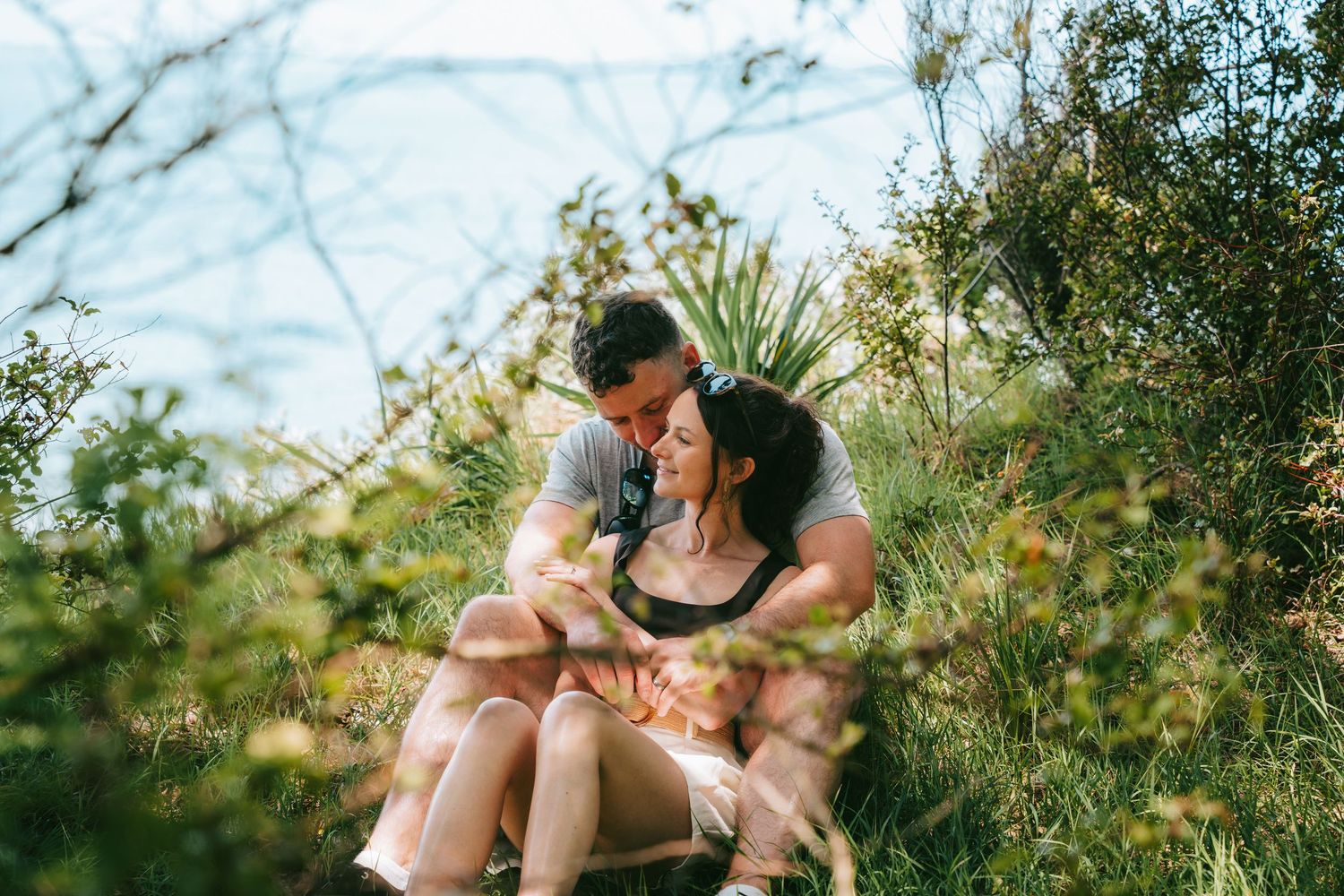 A couple embraces while sitting in a sunlit meadow with tall grass and wildflowers during golden hour.