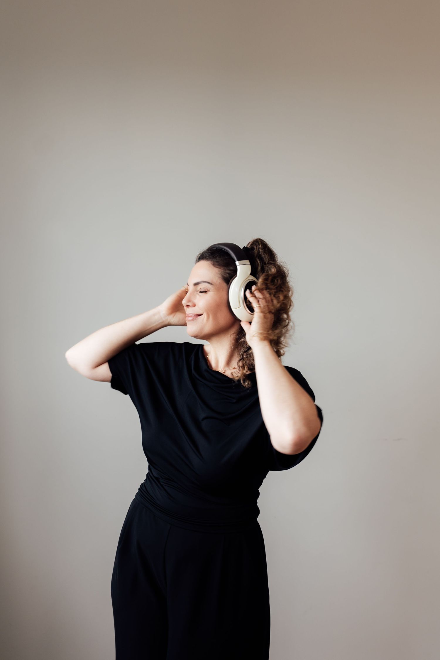 Woman in black outfit enjoying music with white headphones, eyes closed, smiling against neutral background.