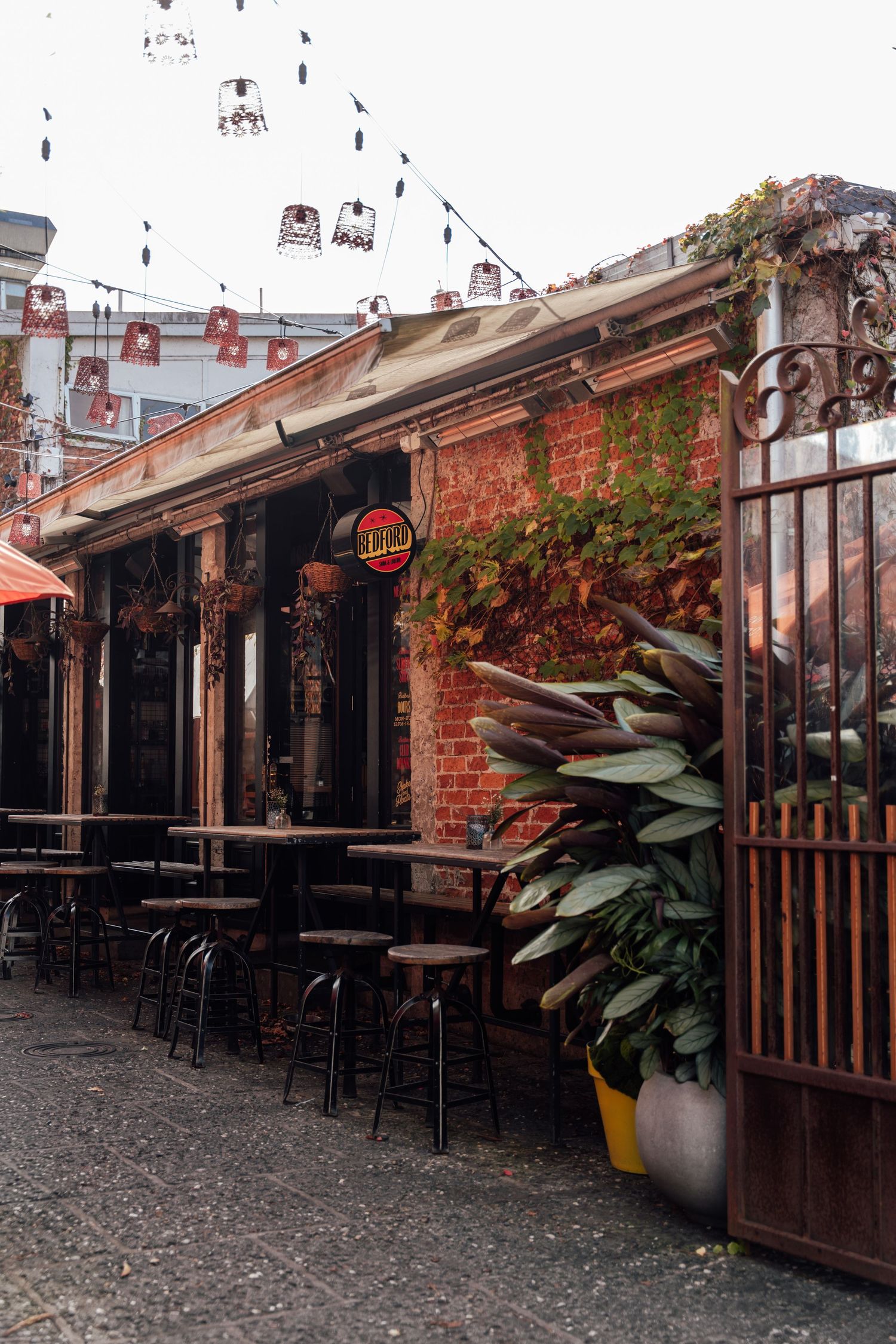 Cozy outdoor cafe patio with black stools, brick walls, hanging lights, and lush green plants on cobblestone street.