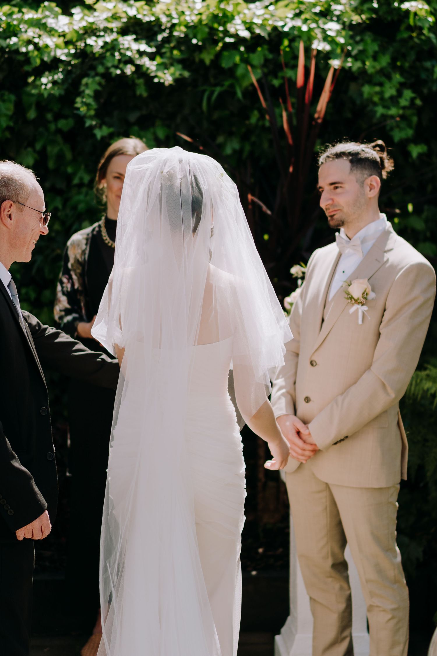 A bride in a white dress and veil stands with guests against a lush green garden backdrop.
