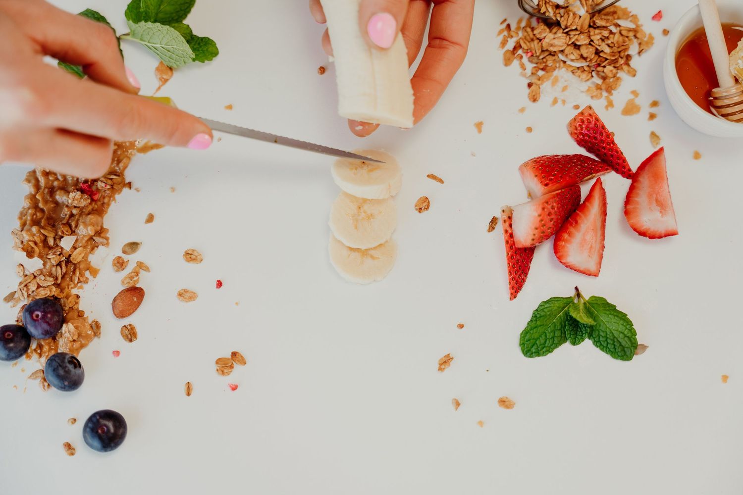 Hands slicing a banana on a white surface surrounded by granola, blueberries, strawberries, and fresh mint leaves.