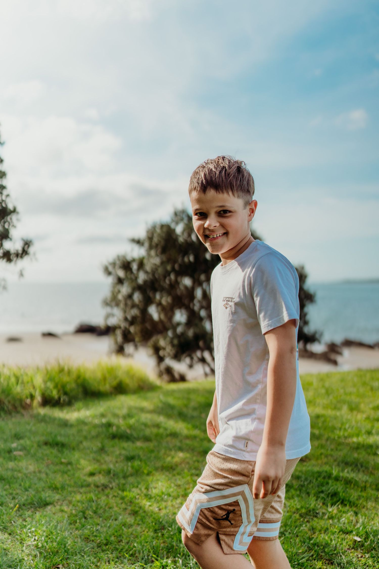 Child in white t-shirt and beige shorts plays on grassy hillside overlooking ocean on sunny summer day.