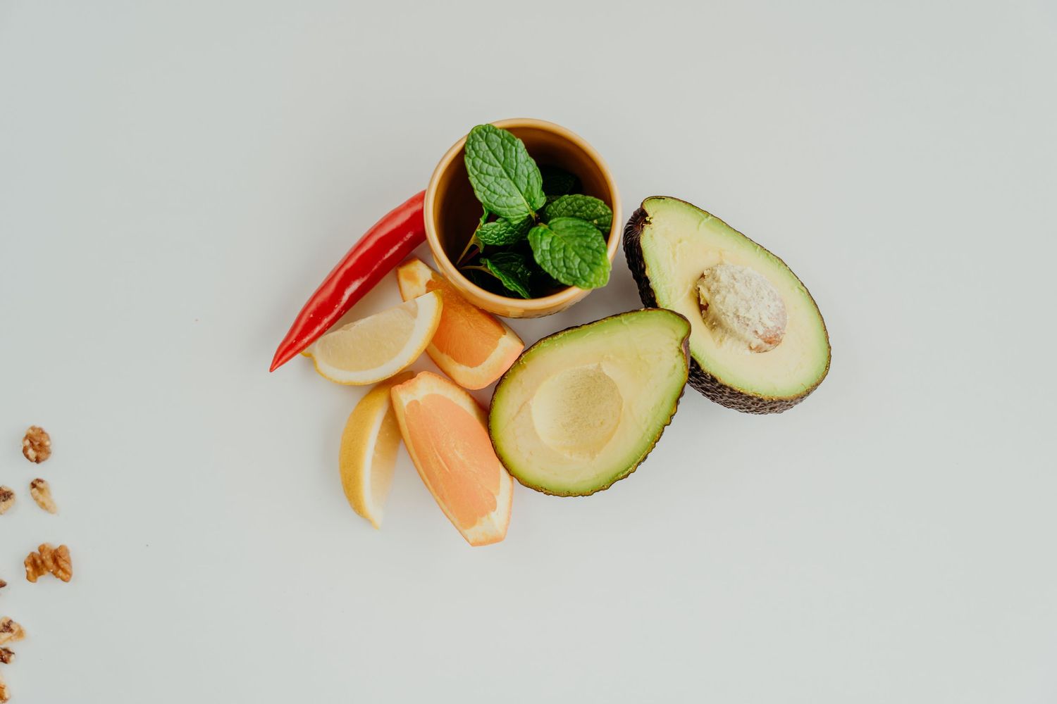 Fresh avocado, citrus slices, red chili, mint leaves, and walnuts arranged on a white background.