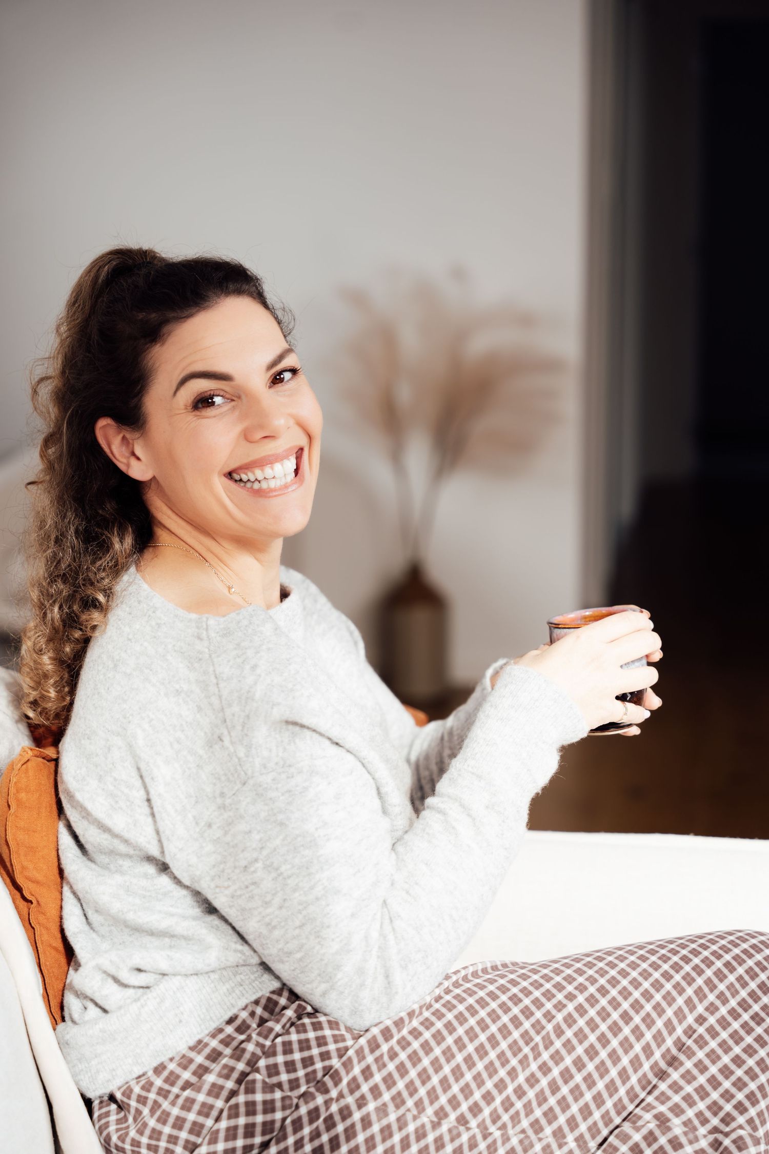Smiling woman relaxing at home in grey sweater holding a coffee mug.
