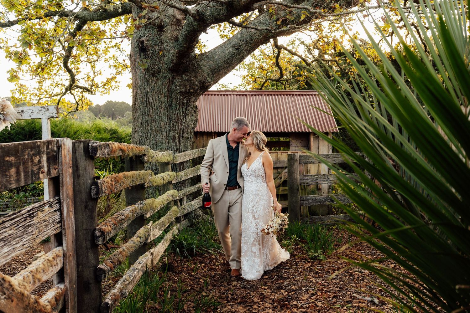 Bride and groom walk hand-in-hand along a rustic fence path beneath a large oak tree at a farm wedding venue.