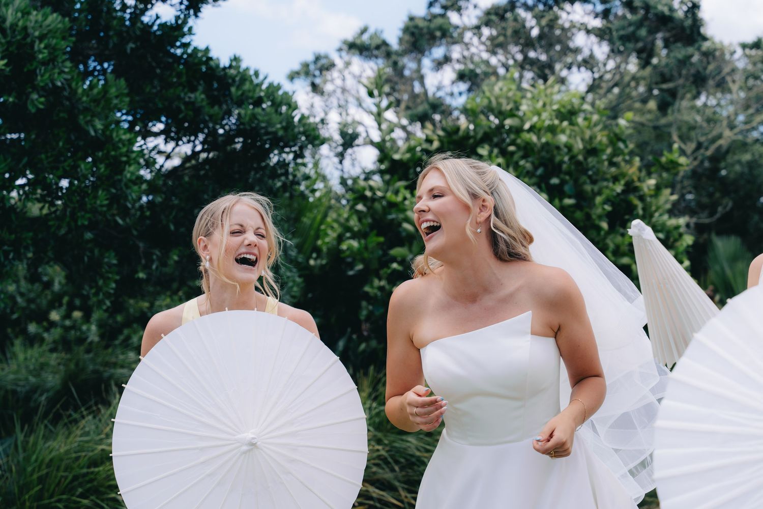 Bride and flower girl laughing joyfully while holding white parasols outdoors at a wedding celebration.