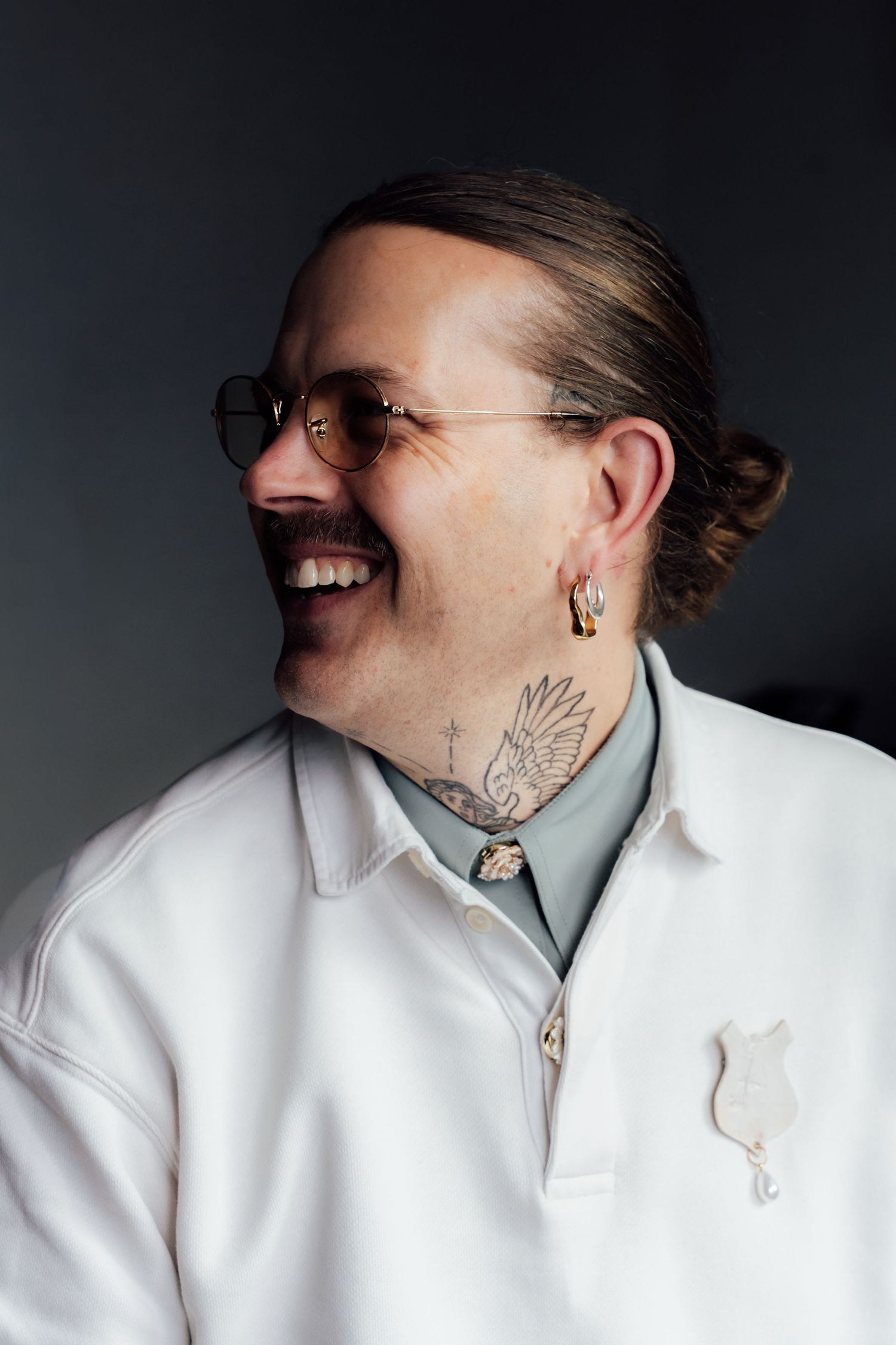 Smiling man with neck tattoo, round glasses, hair bun, gold earring, wearing white jacket and sage green shirt.