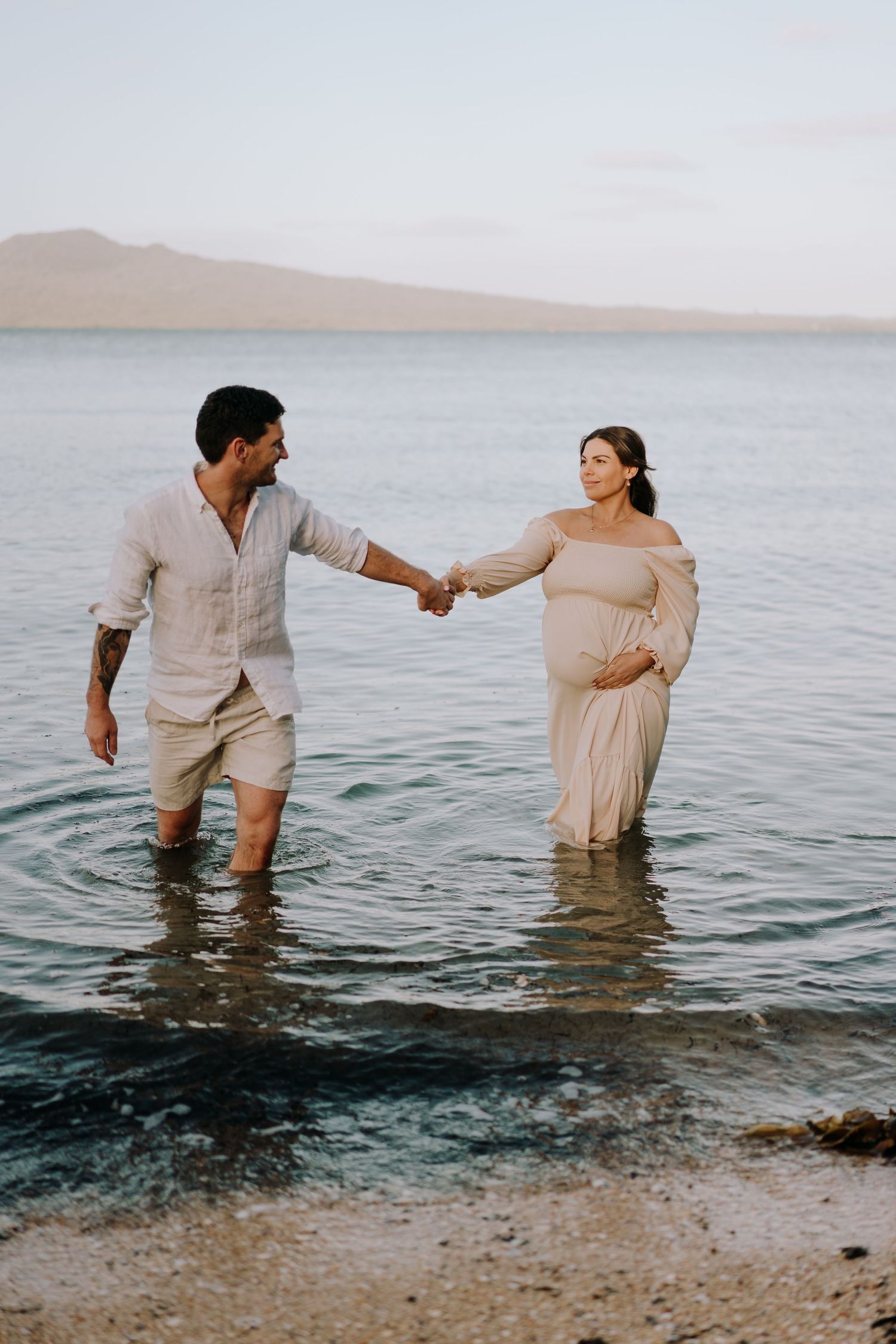 Couple stands in shallow ocean water holding hands during sunset maternity photo session.