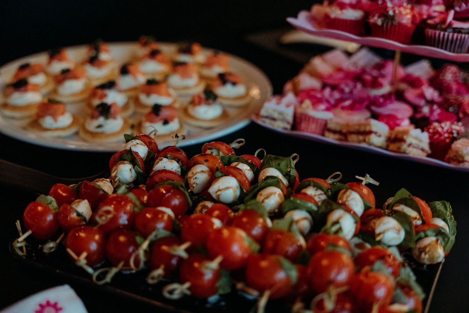Elegant party appetizers display featuring caprese skewers with cherry tomatoes, mozzarella, and pink desserts on a dark buffet table.