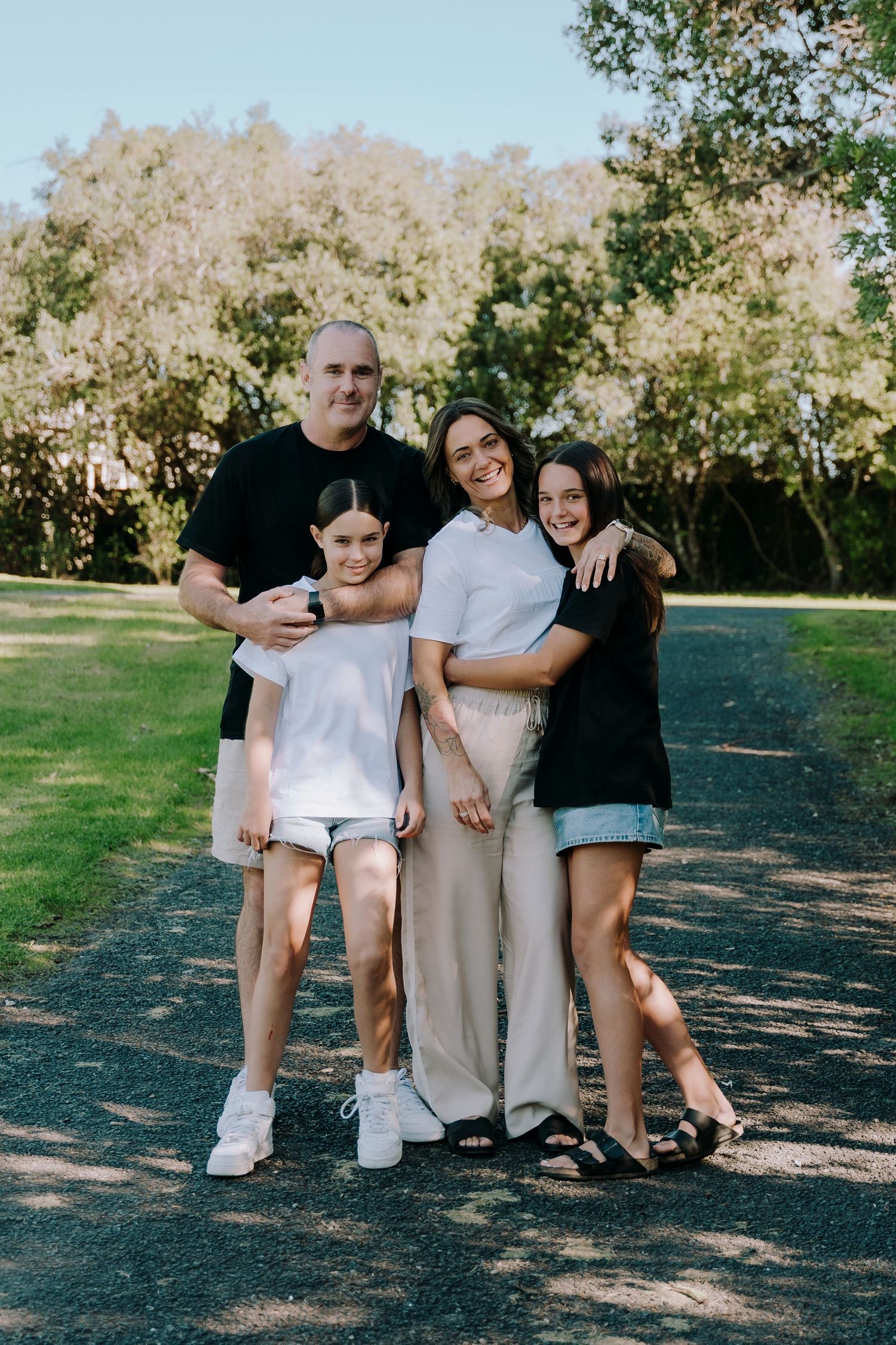 Family group poses together outdoors on a sunny day in a park setting with trees and grass in background.