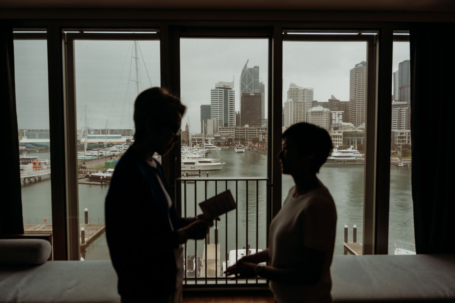 People in silhouette stand at a waterfront railing overlooking a harbor with boats and buildings in the background.