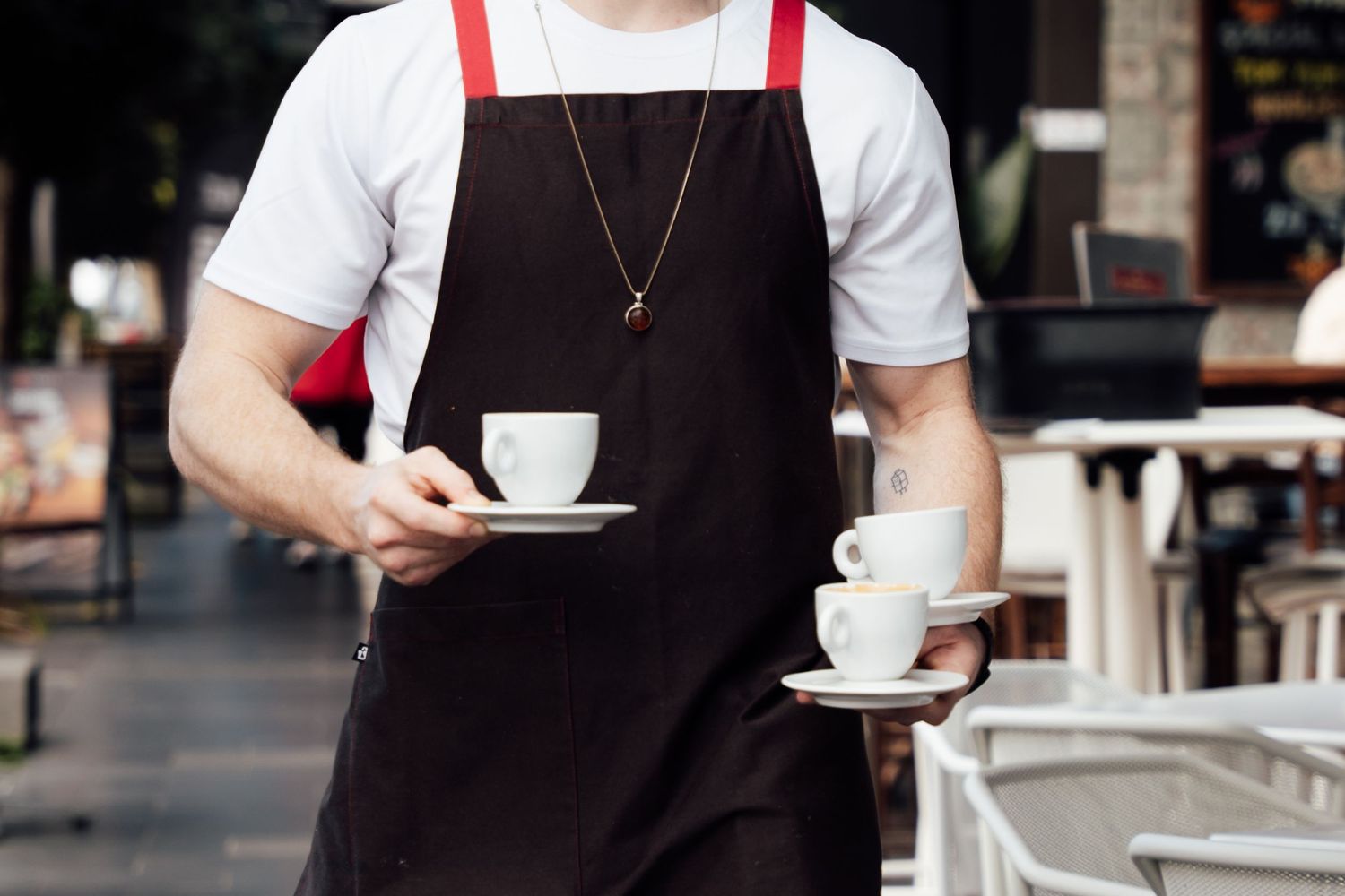 Waiter in dark apron carrying two white coffee cups on saucers at an outdoor cafe.