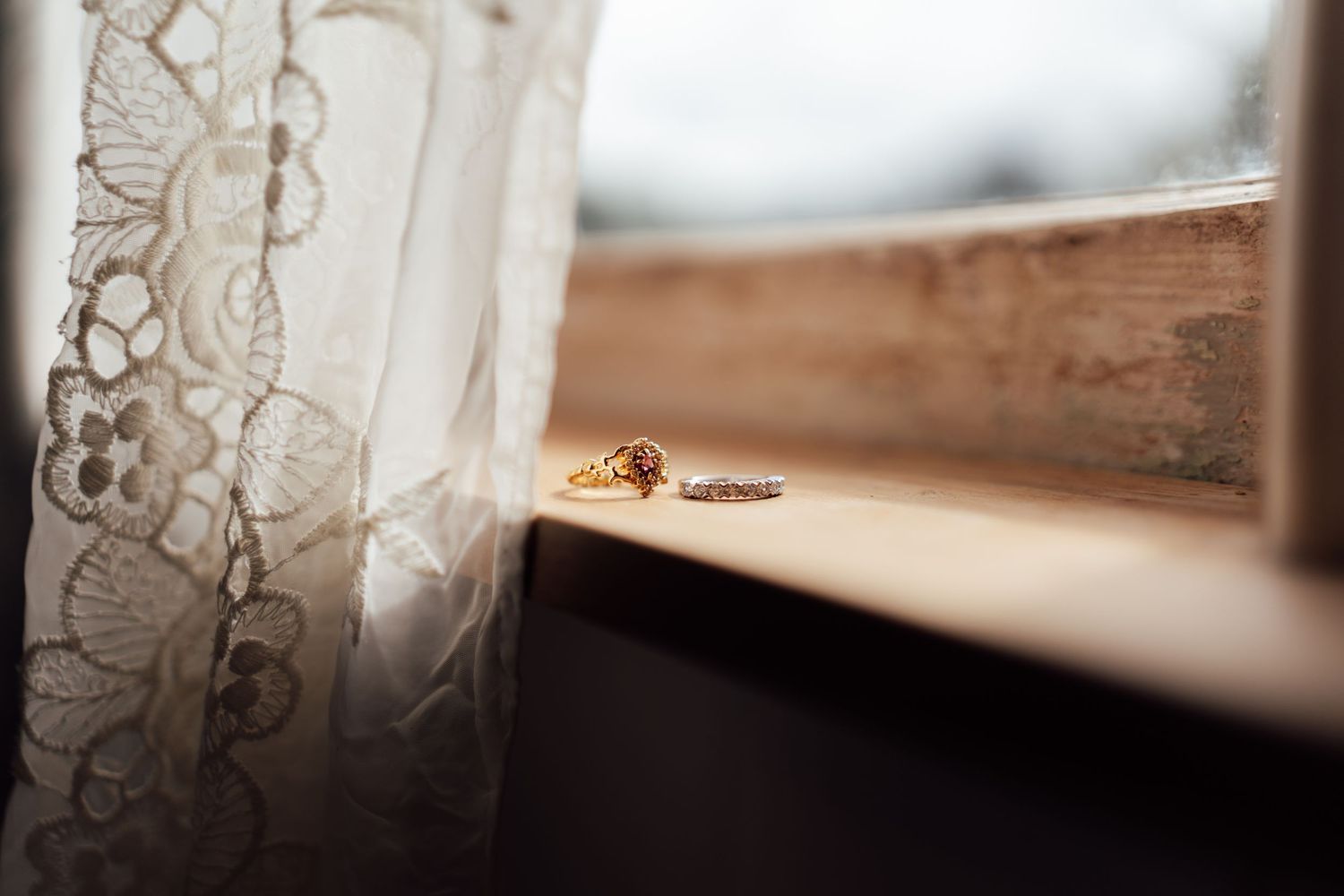 Gold wedding rings resting on a wooden windowsill beside delicate lace curtains in soft natural light.