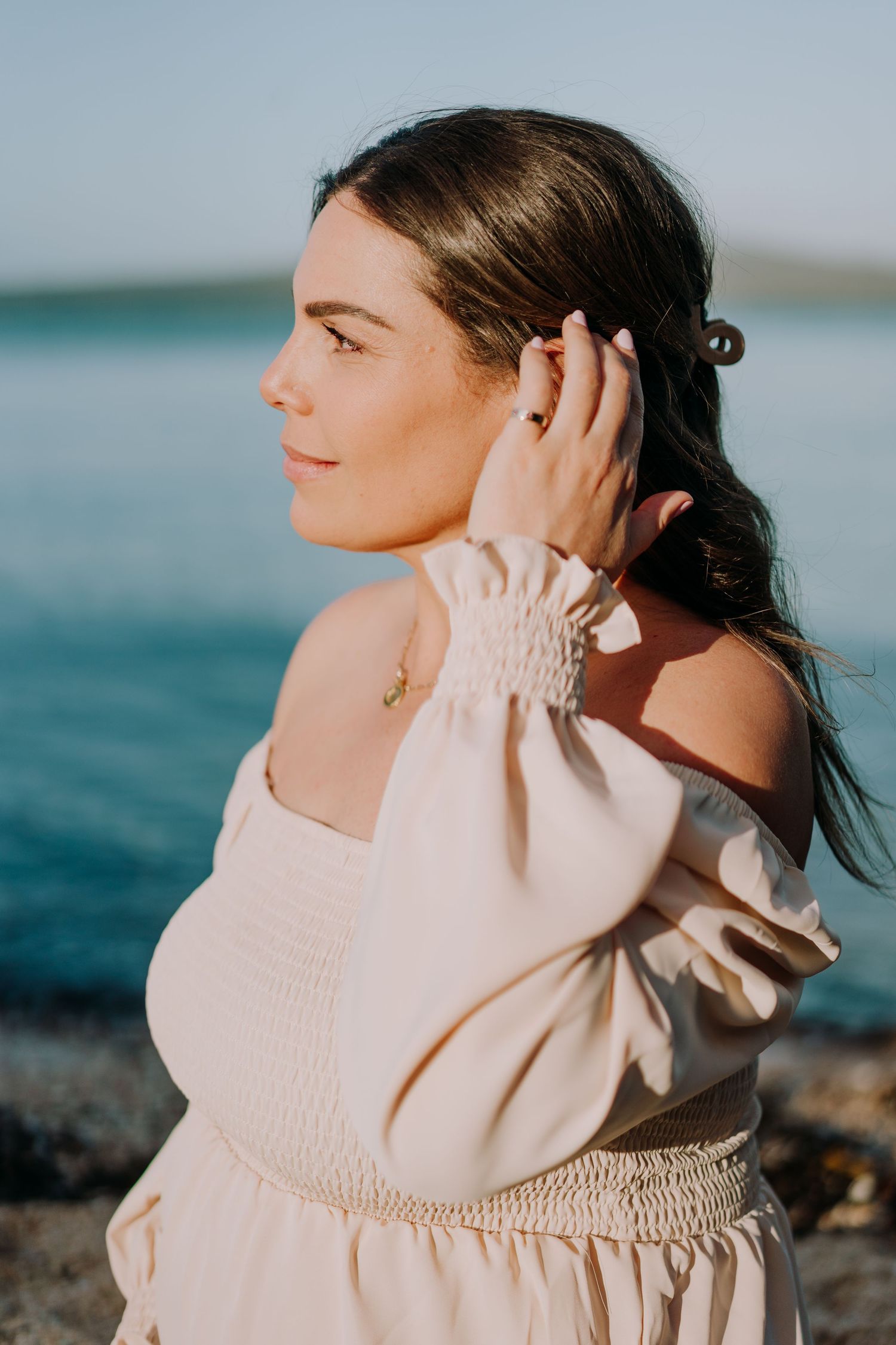 Woman in off-shoulder cream dress poses against turquoise ocean backdrop.