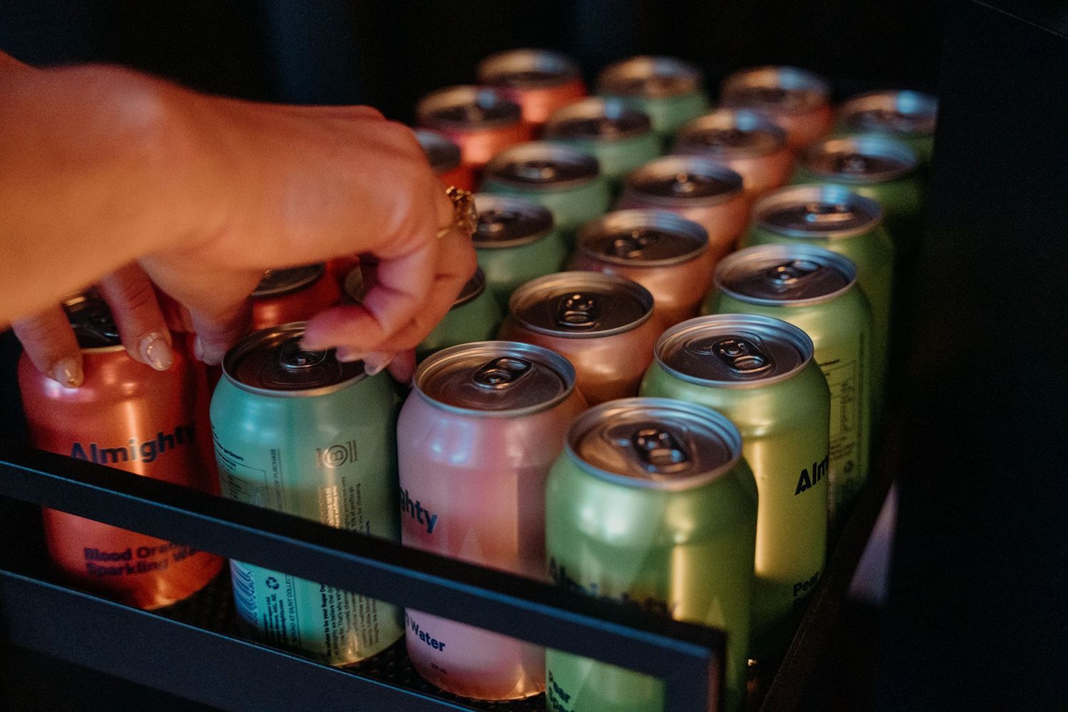 A hand reaching into a refrigerator stocked with colorful aluminum beverage cans arranged in neat rows.