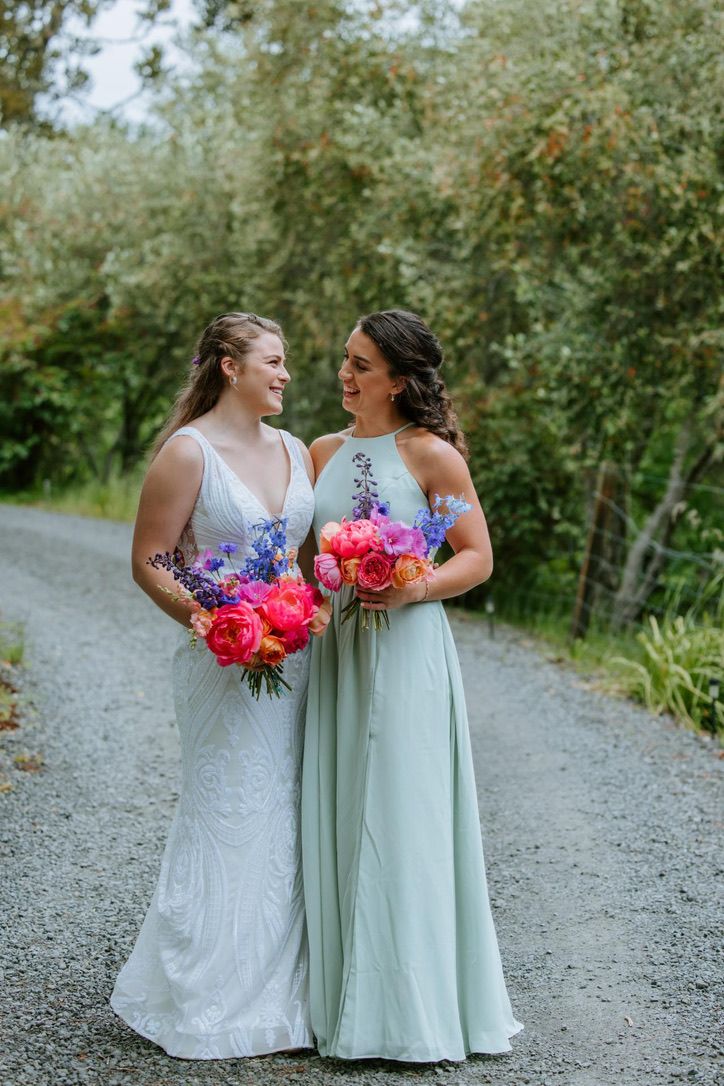 Bridesmaids in white and mint dresses hold vibrant pink bouquets on gravel path.