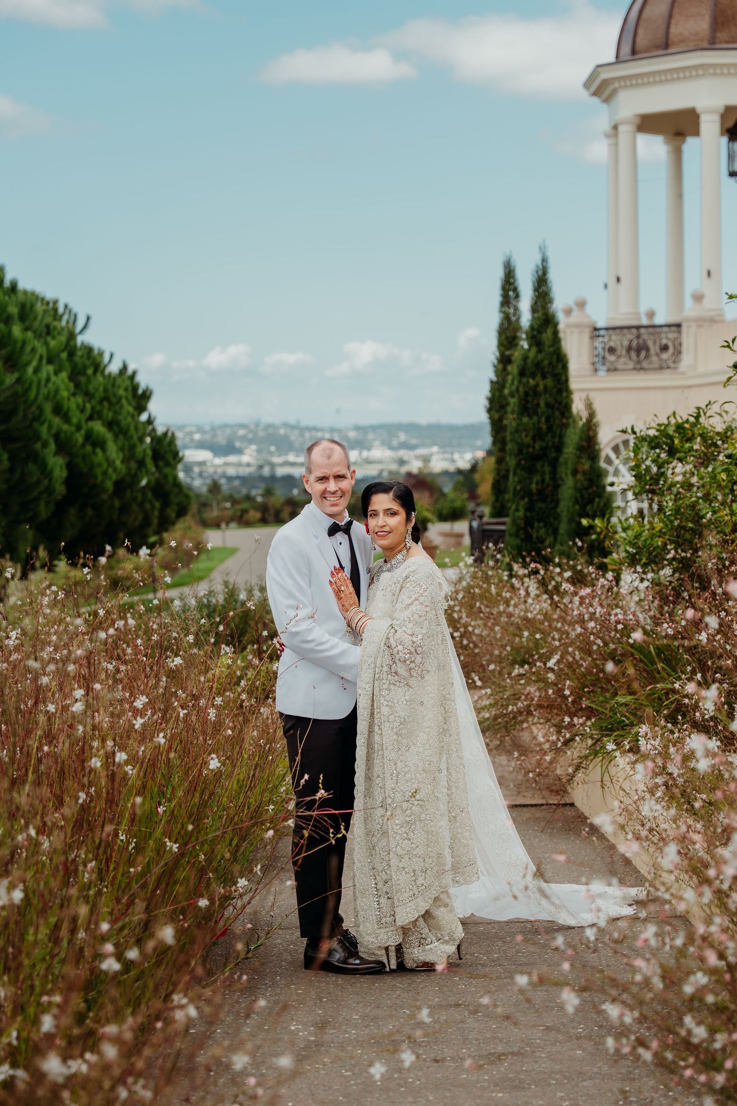 Elegant couple in wedding attire posing in a garden with wildflowers and a white gazebo backdrop.