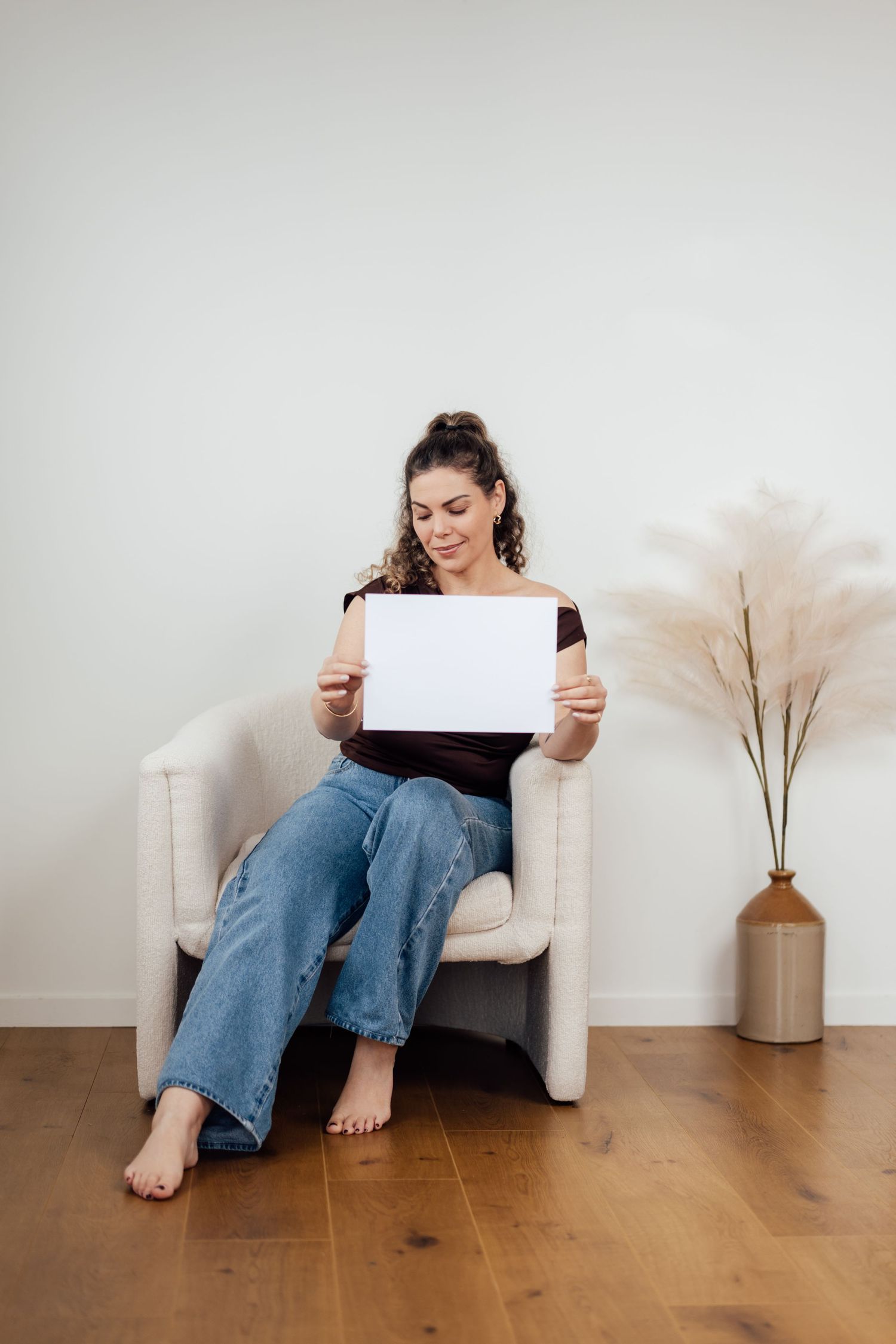Young woman sitting in armchair holding blank white sign, wearing jeans and brown top, barefoot on wood floor.