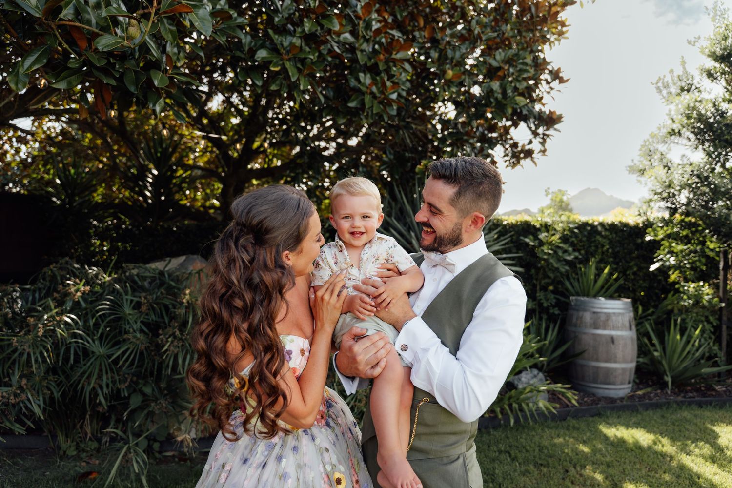 A happy couple in a garden holding their smiling baby, dressed in elegant attire with lush greenery behind them.