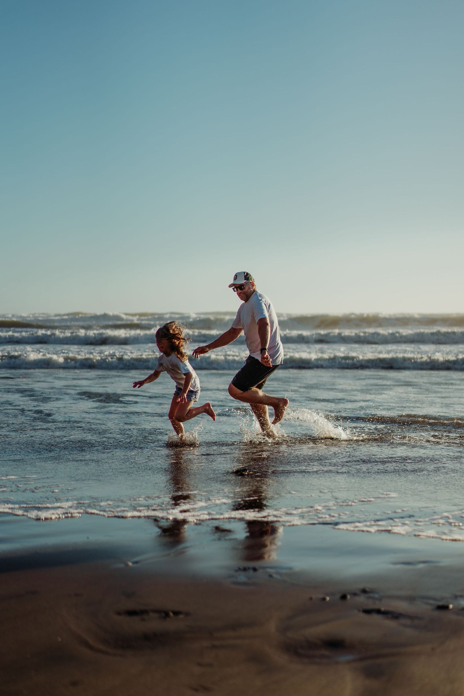 A father and young child running and splashing in ocean waves on a sunny beach with clear blue sky.