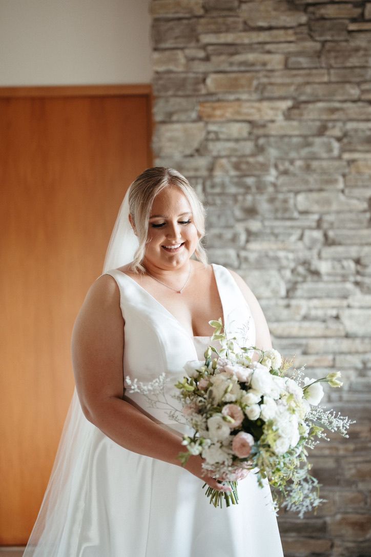 Bride in elegant white gown poses with classic white and green bouquet against stone wall backdrop.