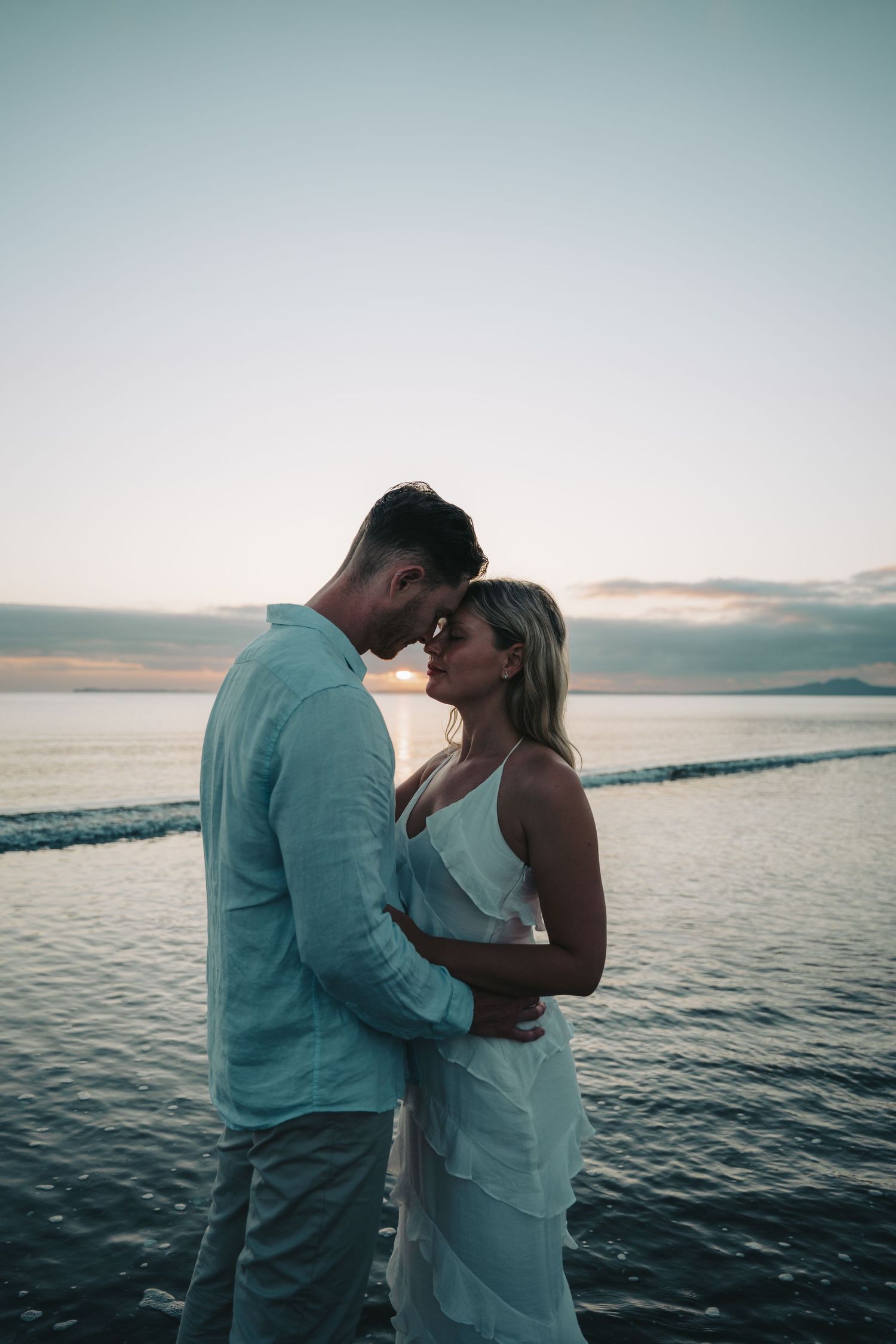 Couple embracing romantically at sunset by the ocean, woman in white dress and man in light blue shirt.