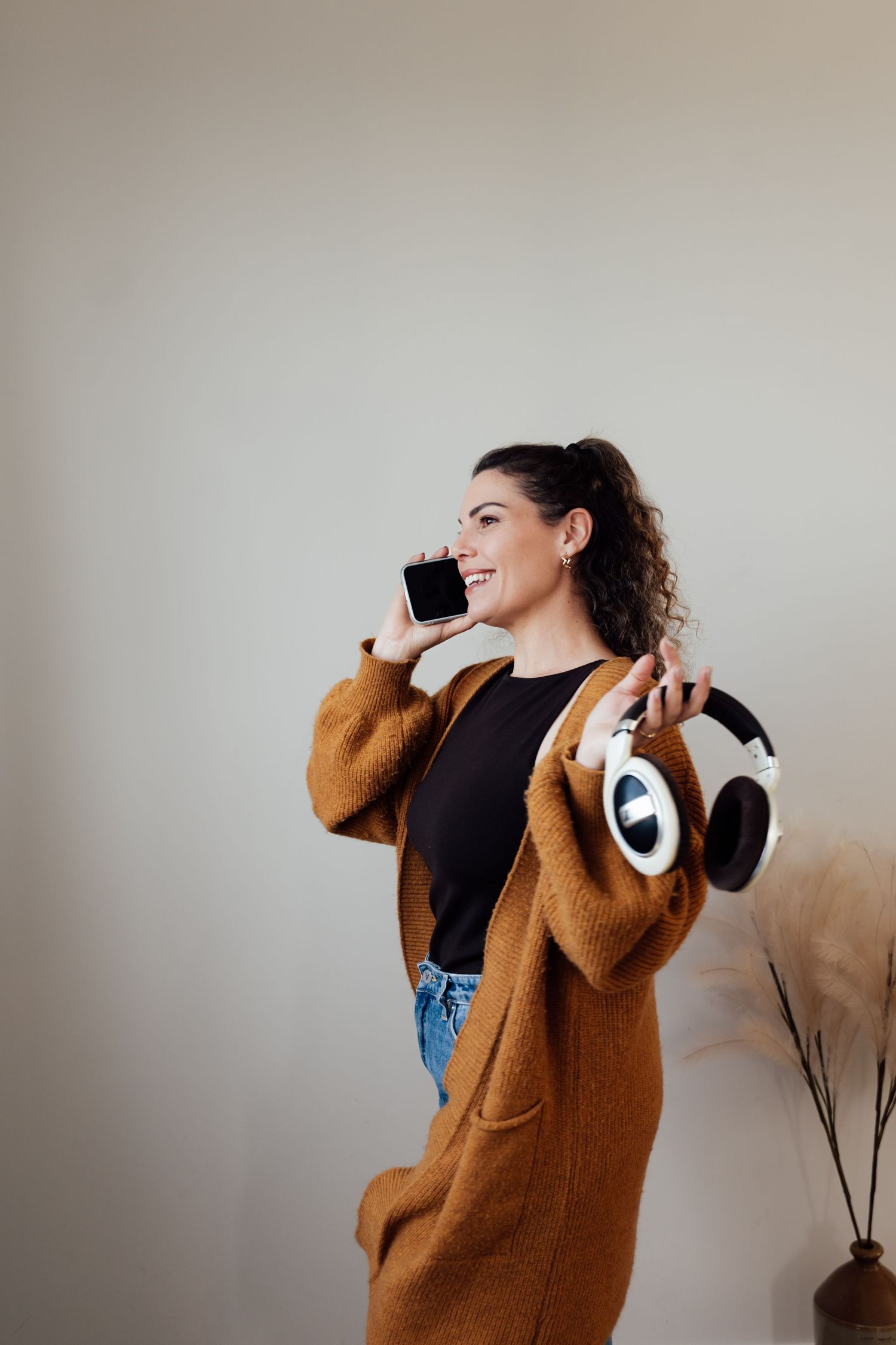 Smiling woman in brown cardigan holding white headphones and smartphone against a light background.