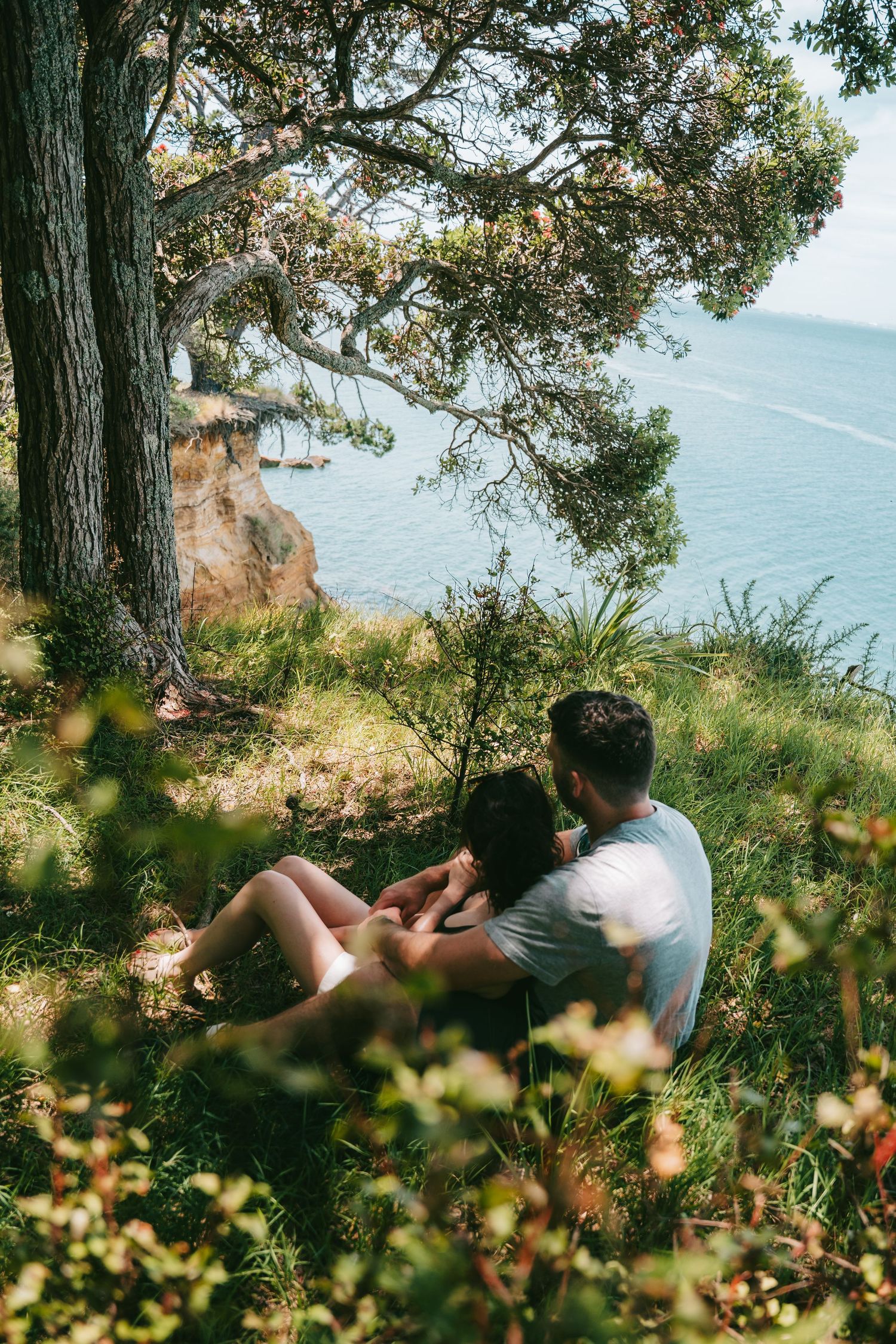A couple sits together overlooking a scenic coastal view through trees and foliage on a sunny day.