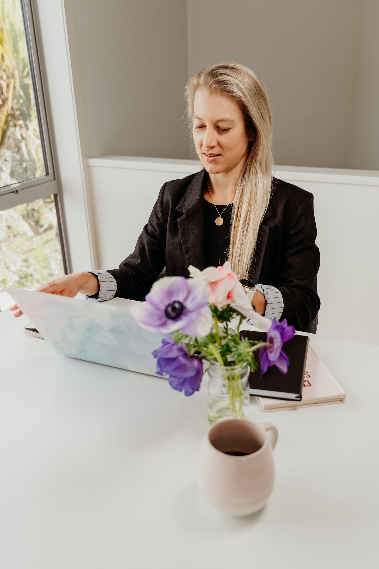 Blonde businesswoman in black blazer working on laptop at white desk with purple flowers and coffee cup.