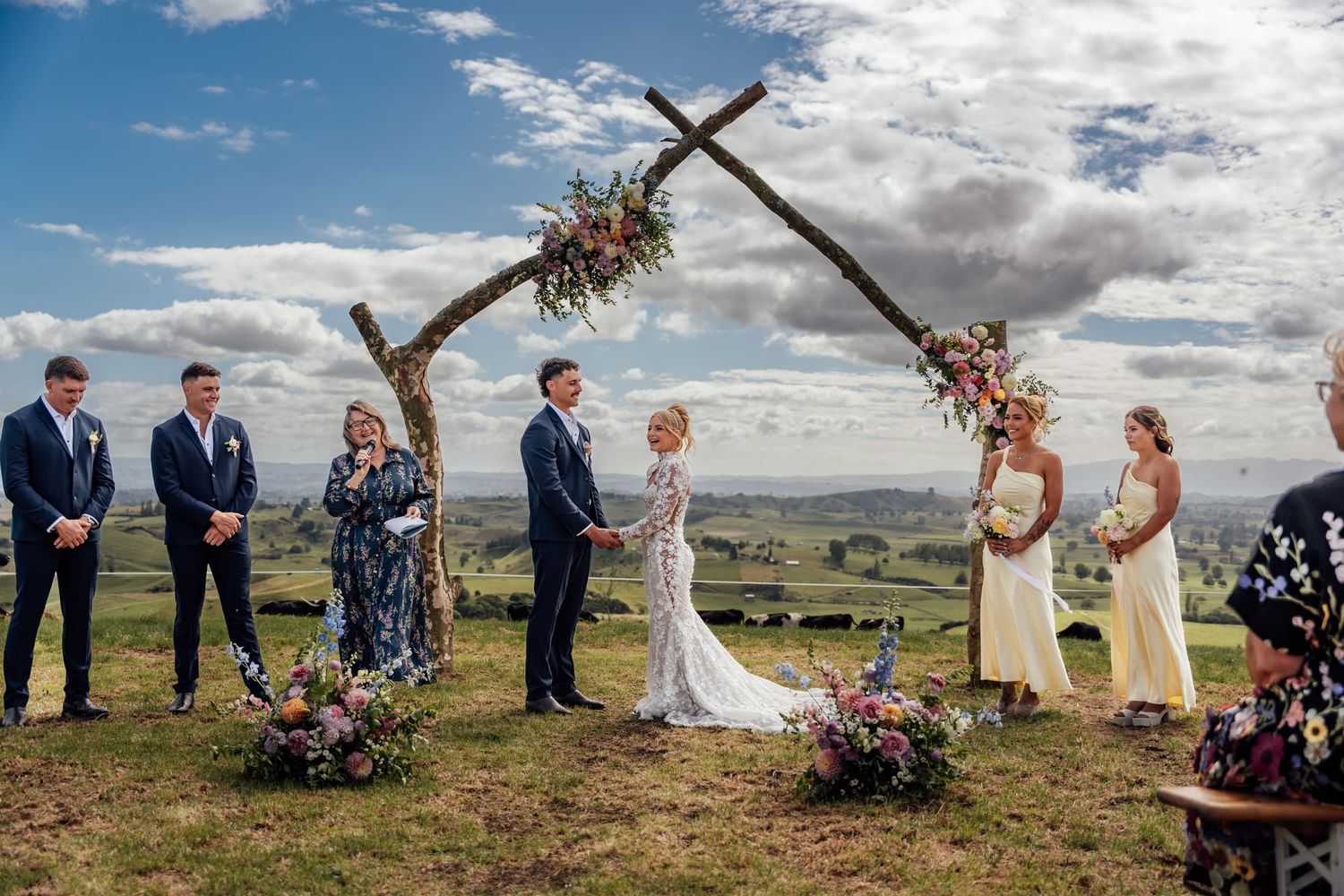 Outdoor wedding ceremony with couple at rustic wooden arch adorned with flowers, overlooking scenic countryside under blue sky.