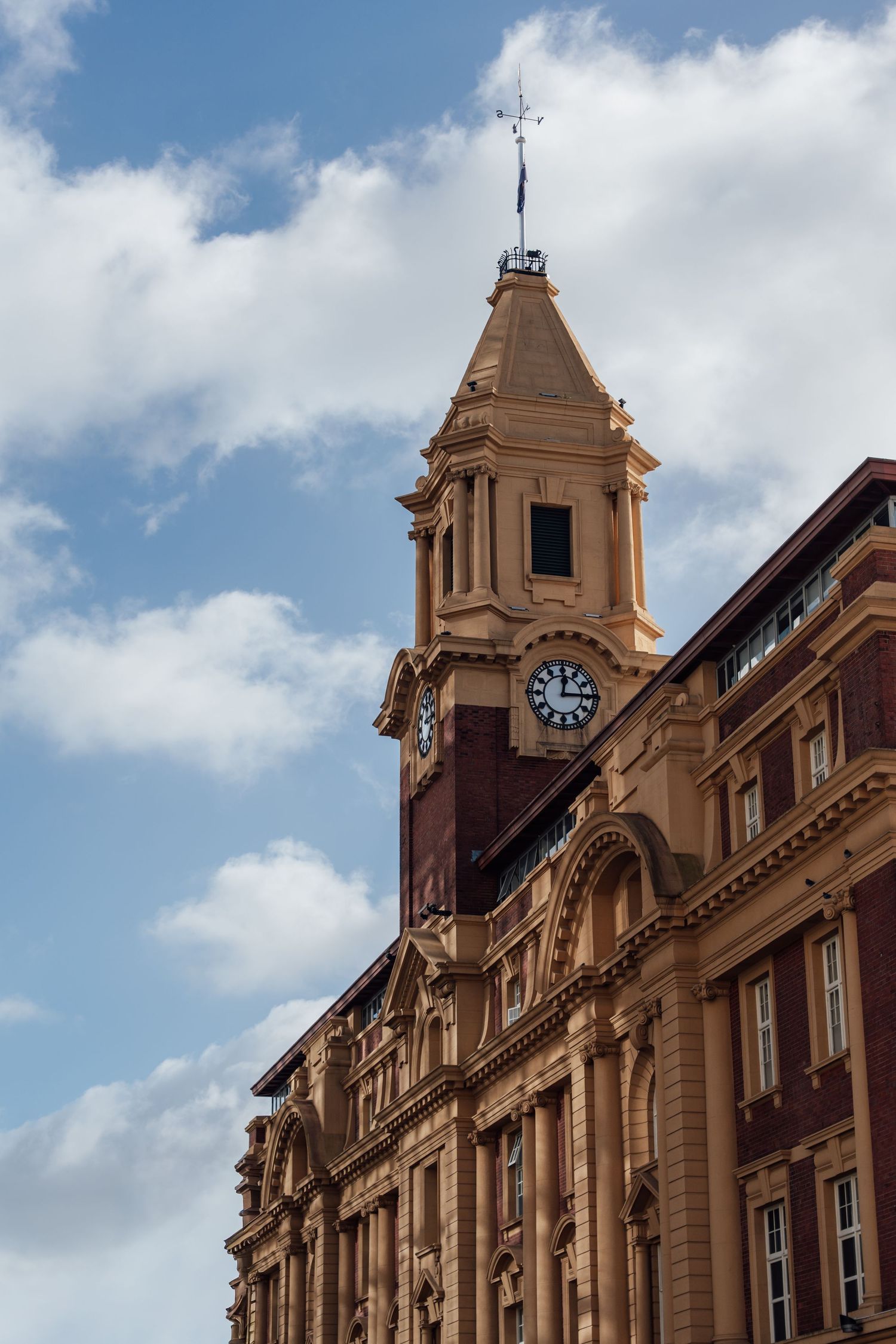 Historic brick building with ornate clock tower and weather vane against a partly cloudy blue sky.