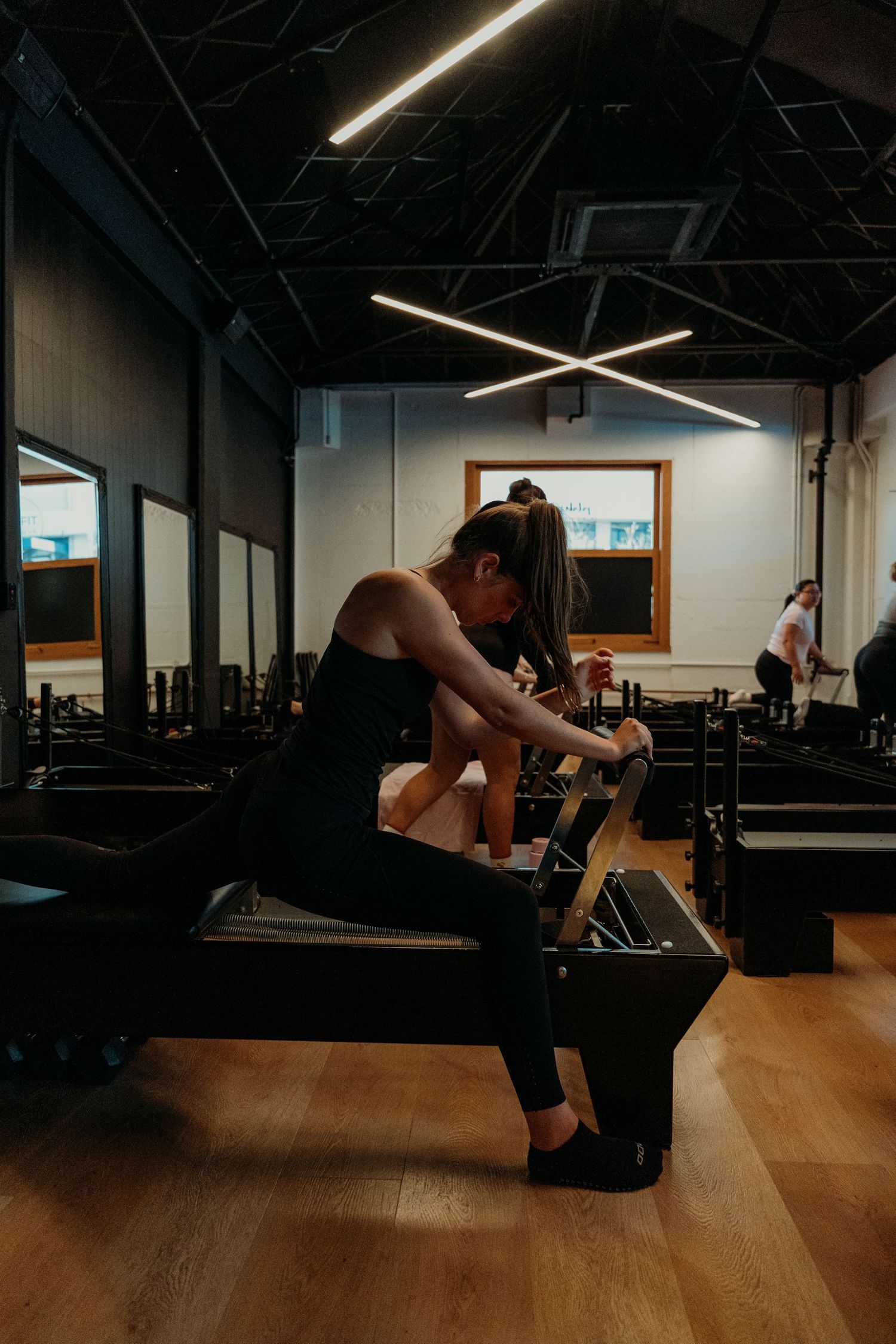 A woman in black activewear using a Pilates reformer machine in a contemporary fitness studio with mirrored walls.