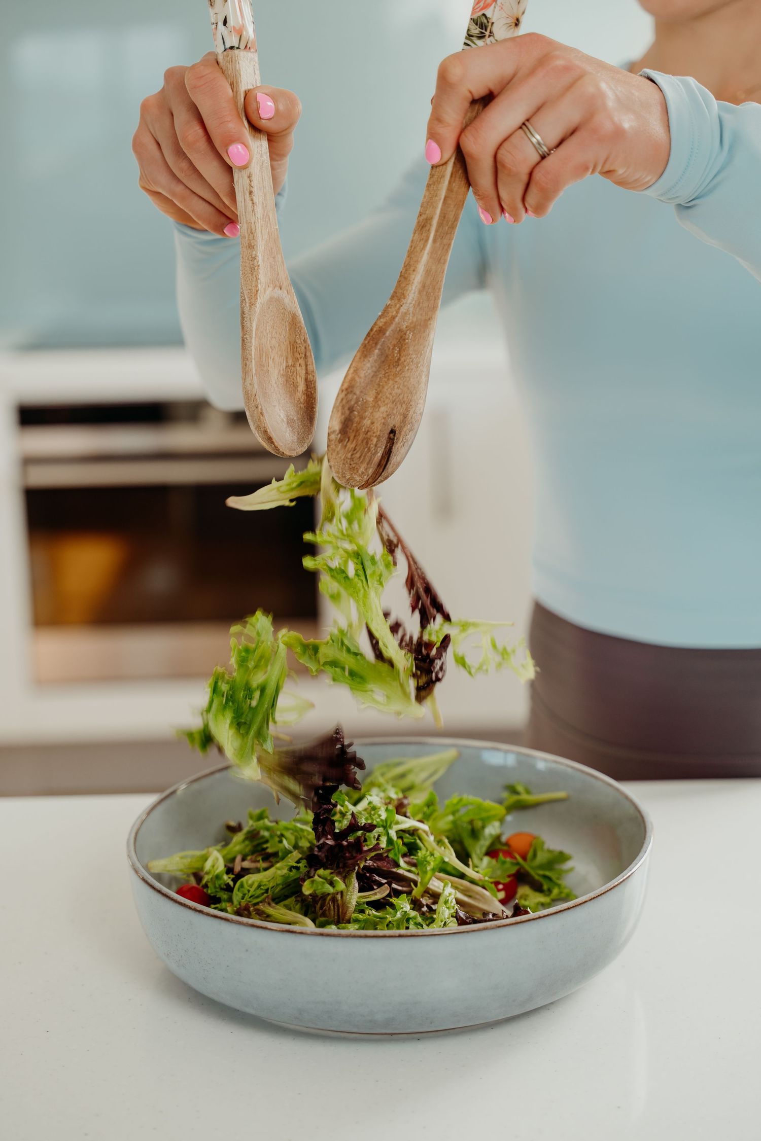 Woman tossing fresh green salad with wooden serving tongs in a modern kitchen bowl.