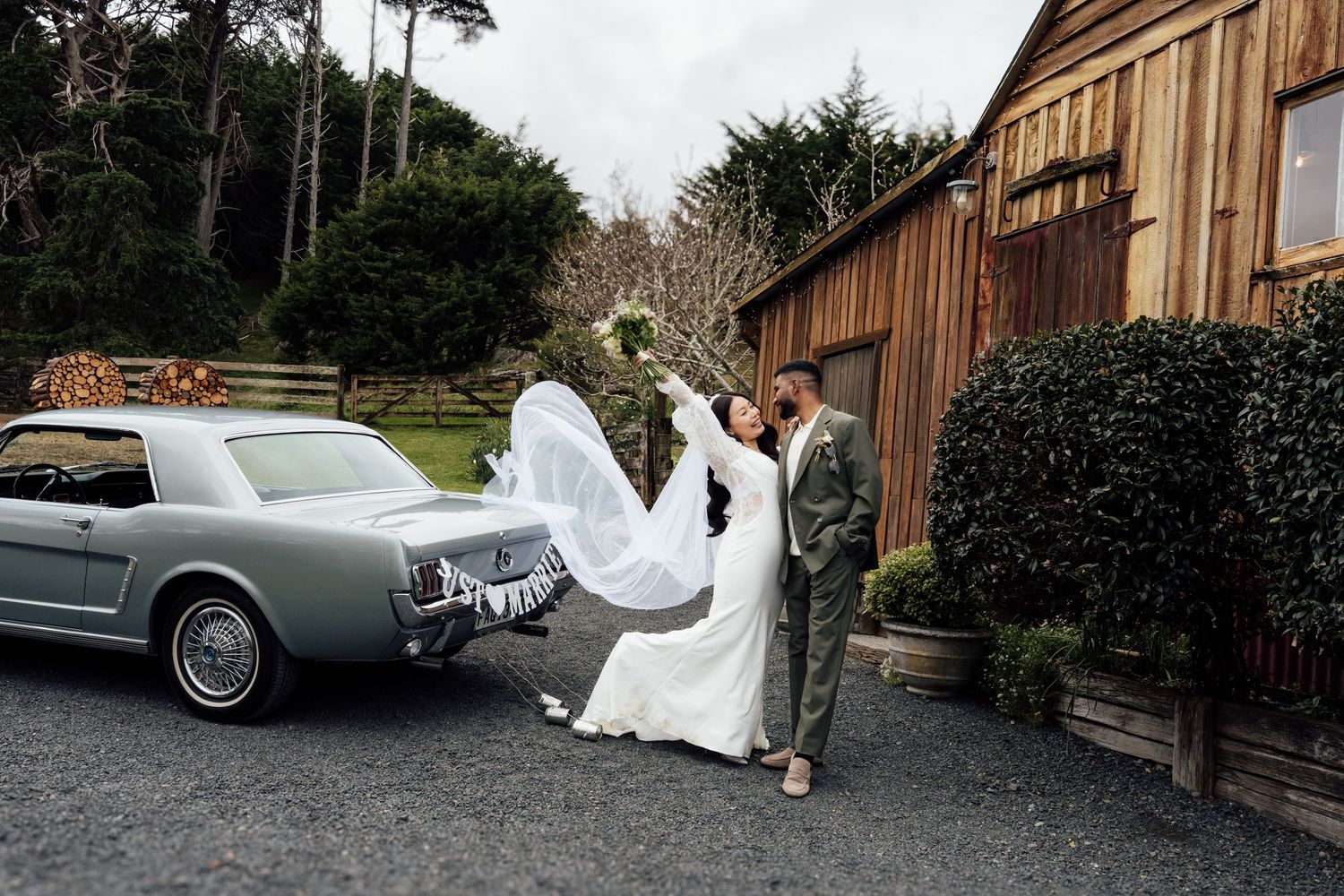 Bride and groom pose beside a classic blue Ford Mustang outside a rustic barn venue, veil flowing in the breeze.