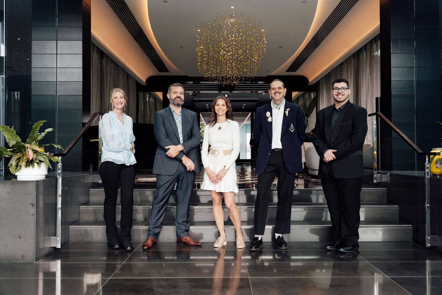 Five professionals posing together in a modern hotel lobby with elegant chandelier and marble floors.