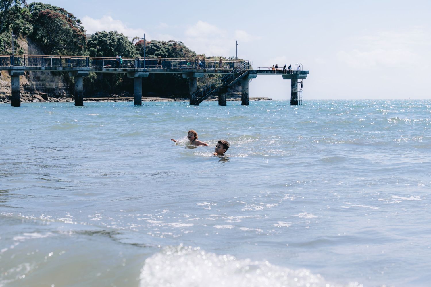 Two children swimming in ocean waves near a wooden pier on a sunny day.