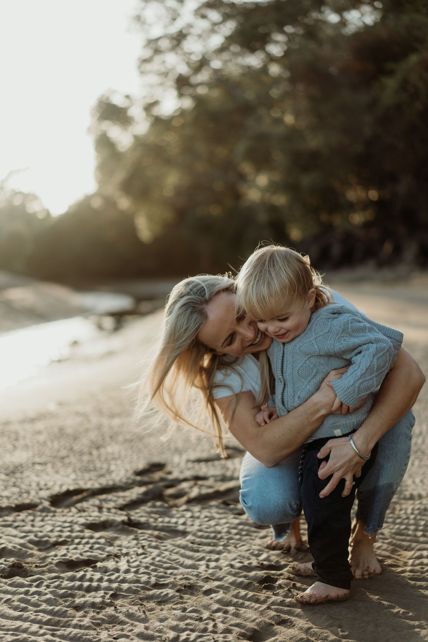 A mother hugs her young blonde child on a sandy beach at golden hour with trees in the background.