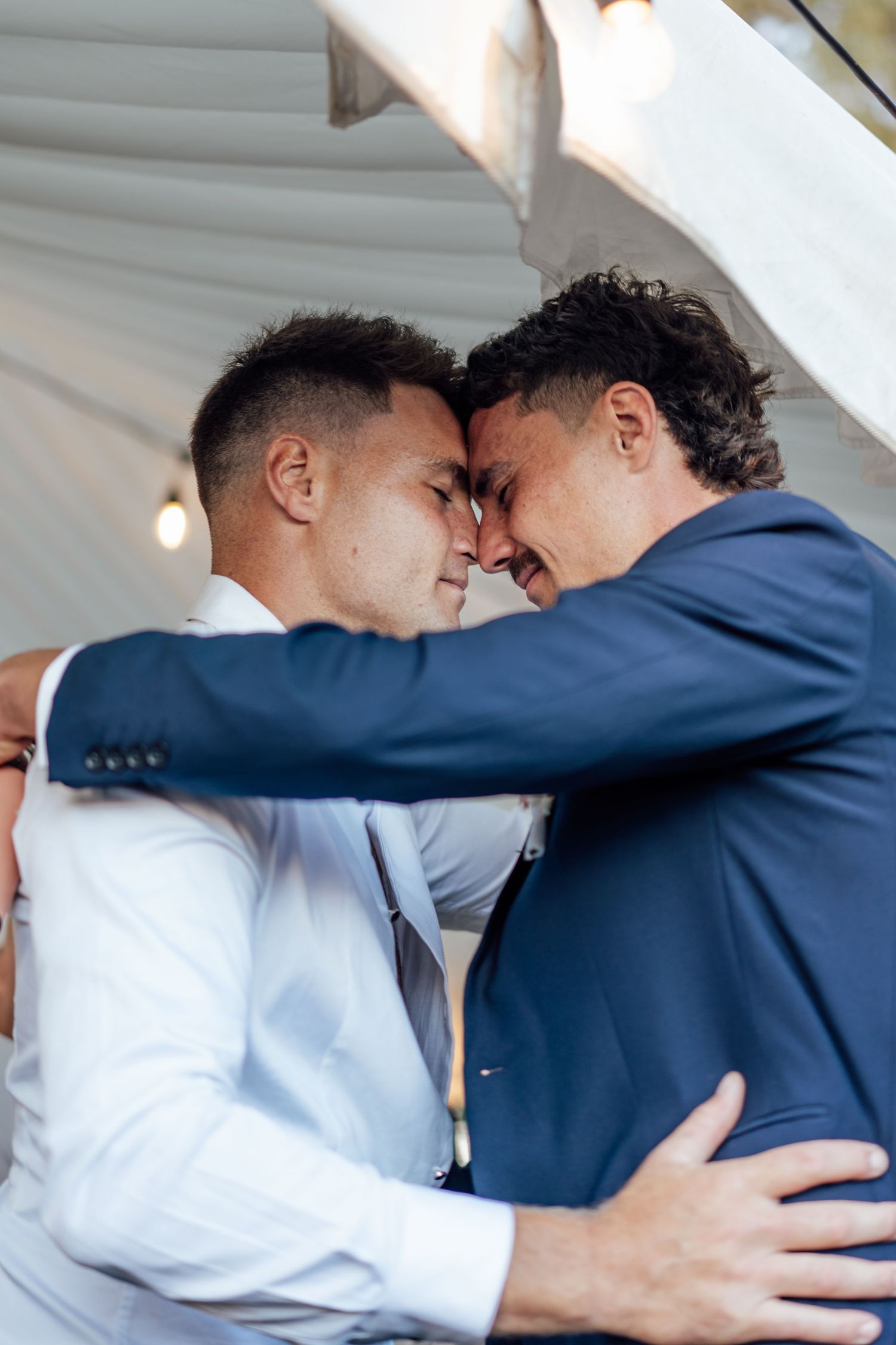 Two grooms in suits embrace tenderly, touching foreheads in an intimate wedding moment.