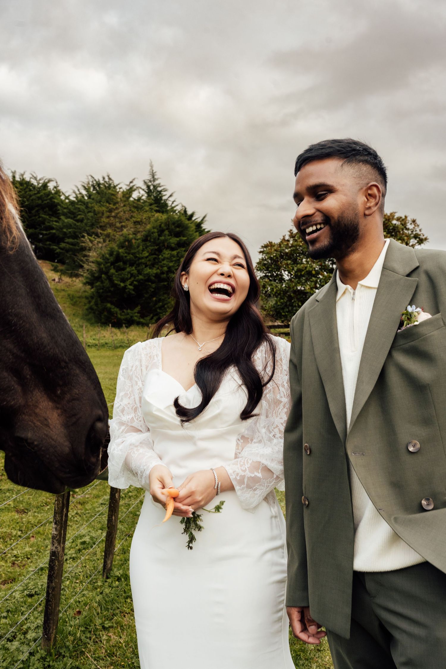 Happy interracial couple in wedding attire laughing beside a horse in a lush green field on their wedding day.