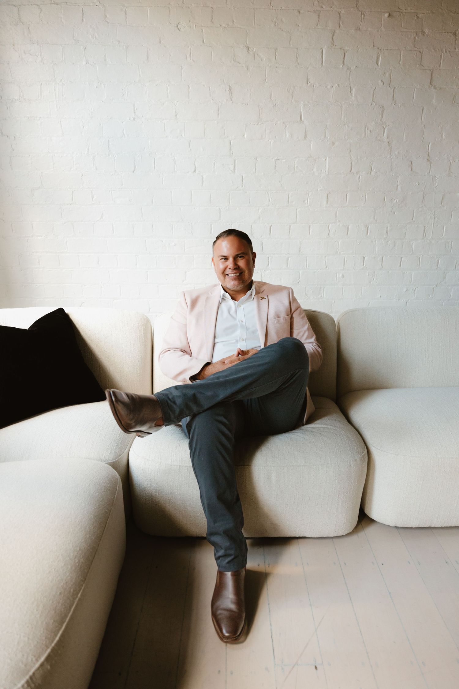 Smiling man in pink blazer and gray pants sitting relaxed on a cream sofa against a white brick wall.
