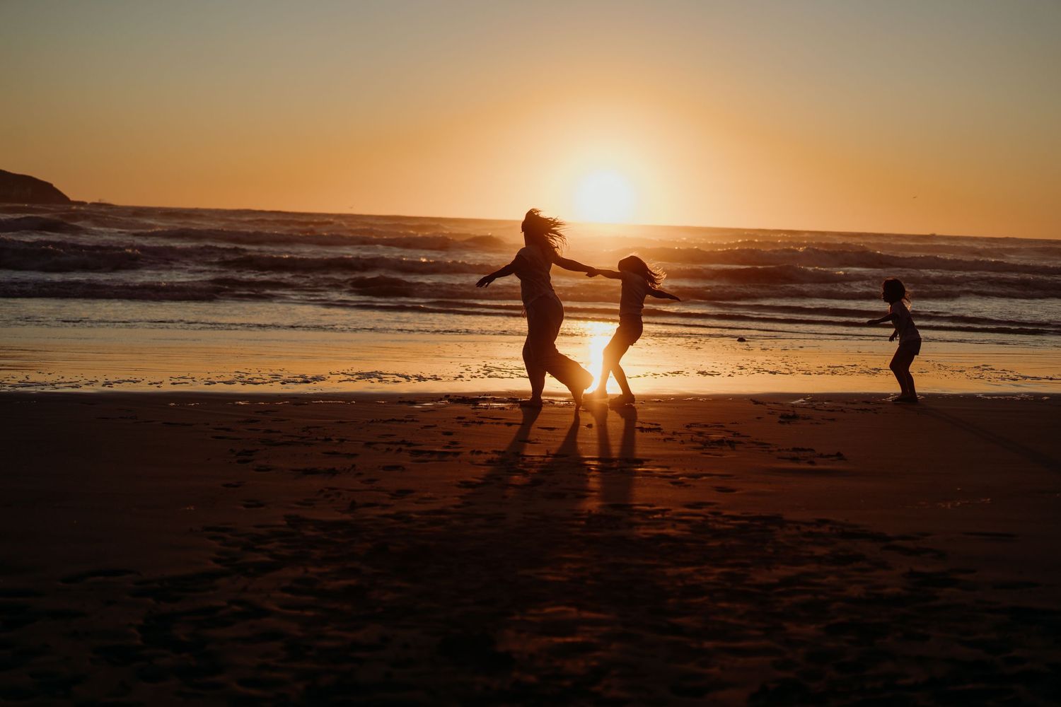 Silhouettes of a mother and two children playing on a beach at golden sunset with ocean waves in the background.