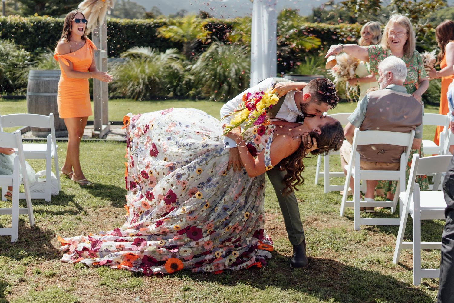 Couple shares romantic dip kiss at outdoor wedding ceremony, bride in floral gown, guests seated on white chairs.