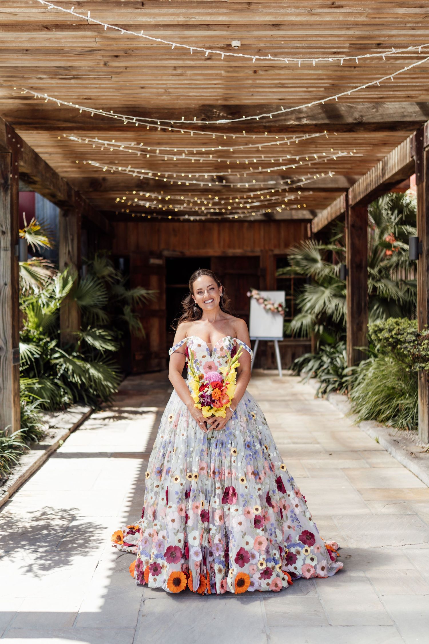 Bride in colorful floral off-shoulder ball gown holding vibrant bouquet under string lights at rustic outdoor venue.